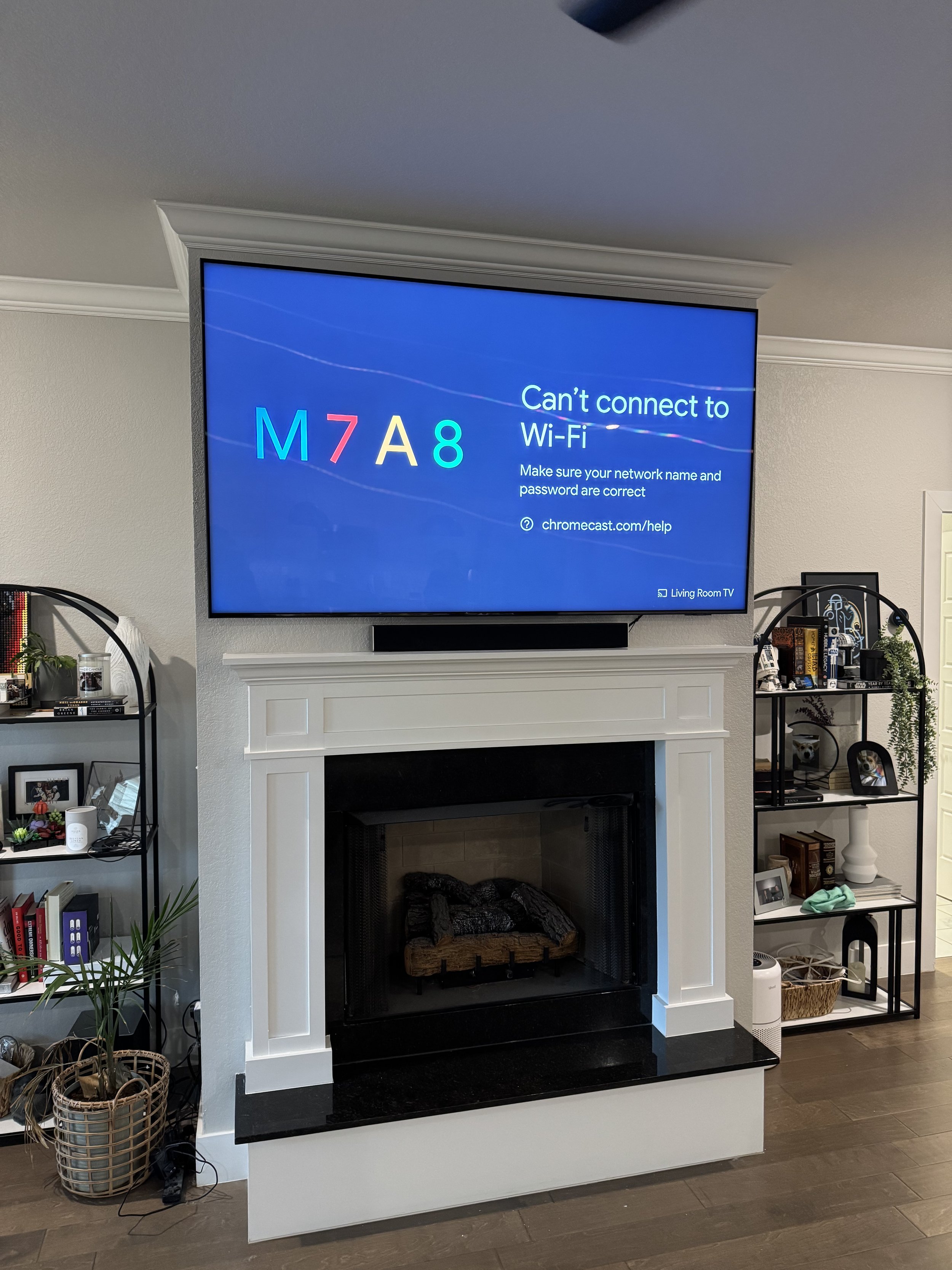 A living room with a mounted flat-screen TV displaying a Wi-Fi connection error message, above a white fireplace with black accents, flanked by black metal shelves filled with books, decorative items, and framed photos, on a dark wood floor.