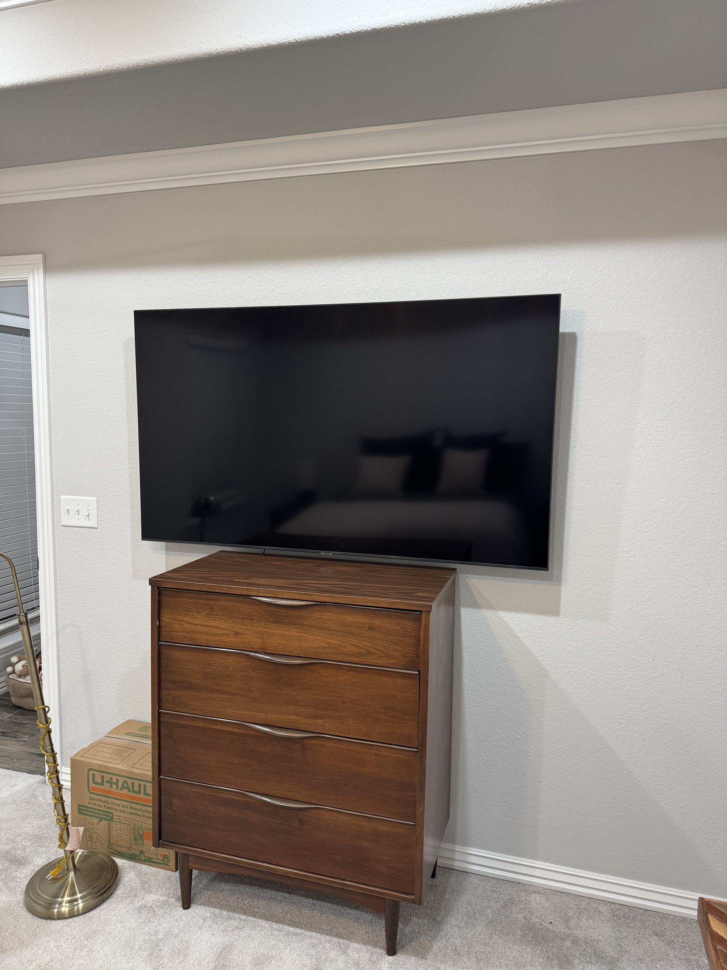 A flat-screen TV mounted on a white wall above a tall wooden dresser in a living room. A U-Haul box and a gold-colored floor lamp are visible nearby.