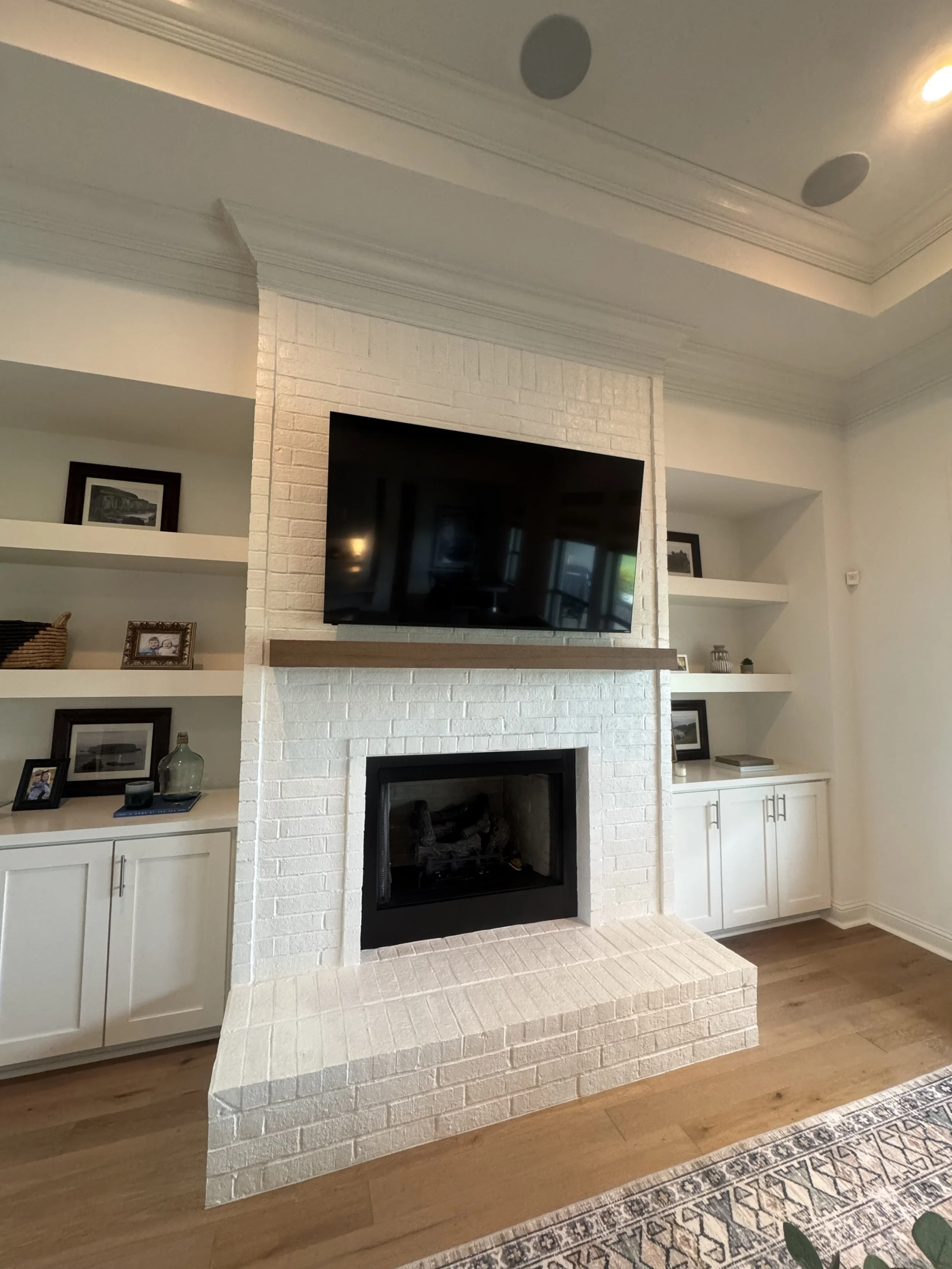 Living room fireplace with a mounted flat-screen TV above it, surrounded by built-in white shelves with framed photos, decorative objects, and books.