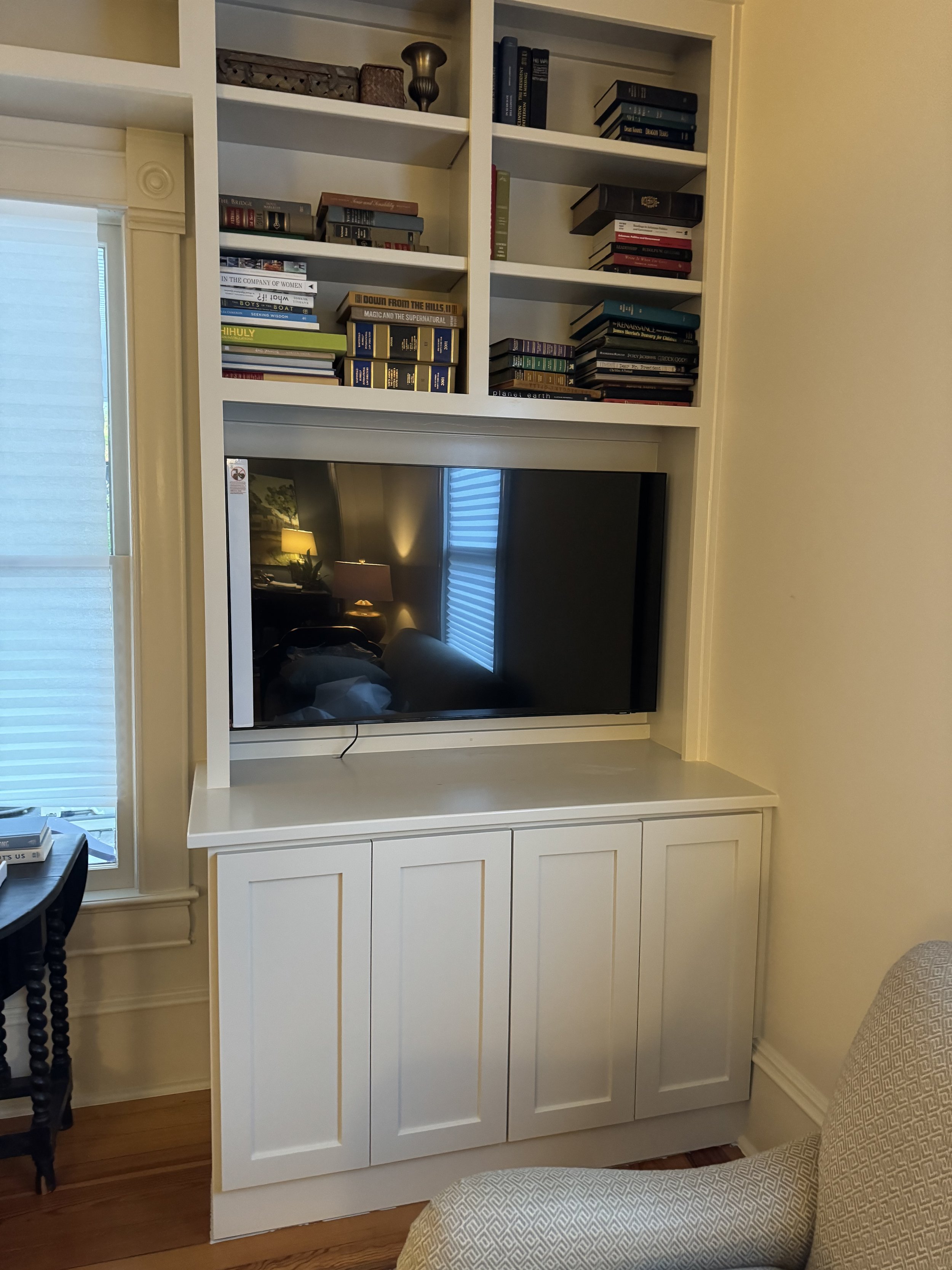 A white built-in entertainment center with bookshelves, a flat-screen TV, and closed cabinet doors in a living room with hardwood floors. Reflection of a lamp and window in the TV screen.
