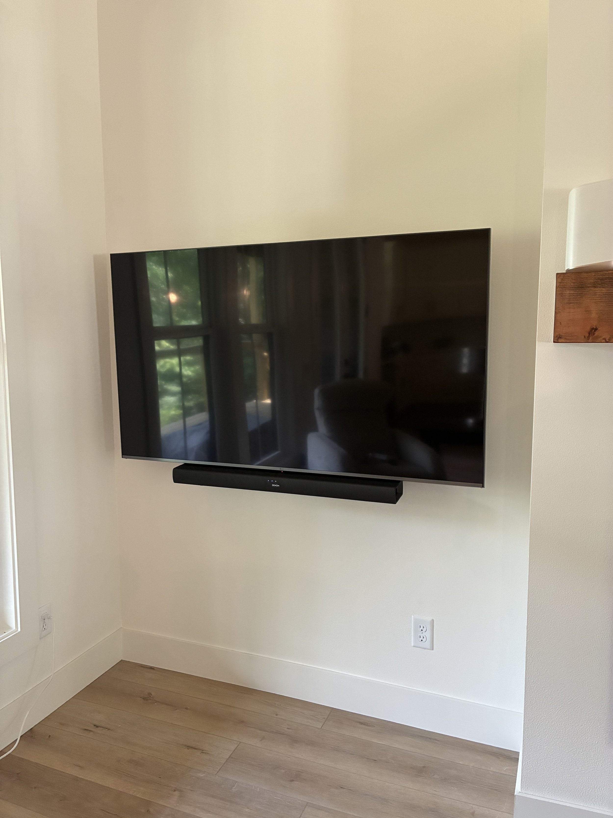 Wall-mounted flat-screen television with a soundbar below, switched off, in a room with light-colored walls and wooden flooring.
