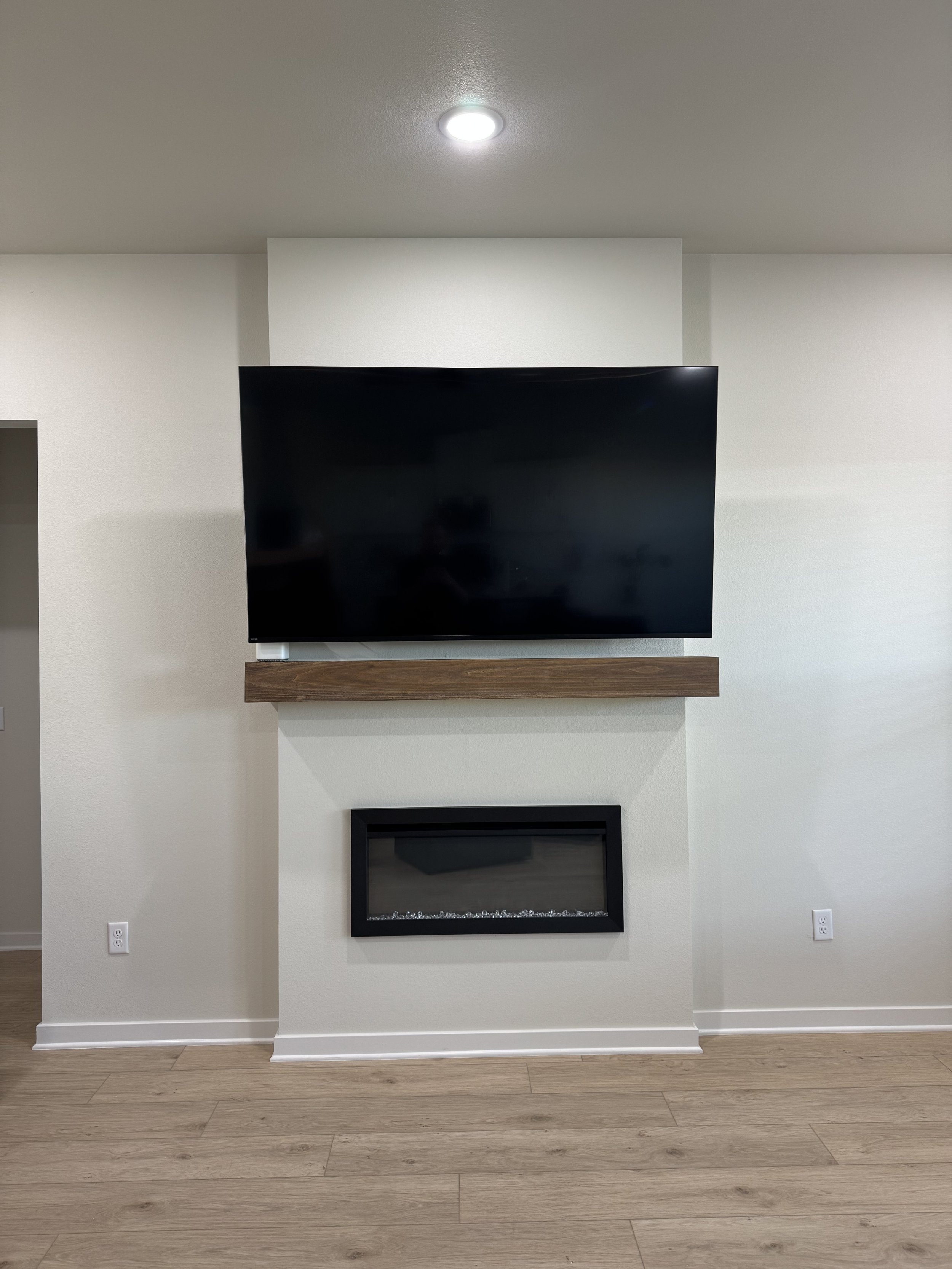 Living room with a mounted flat-screen TV above a modern fireplace with a black frame and decorative stones, and a wooden mantel on a white wall with light wood flooring.