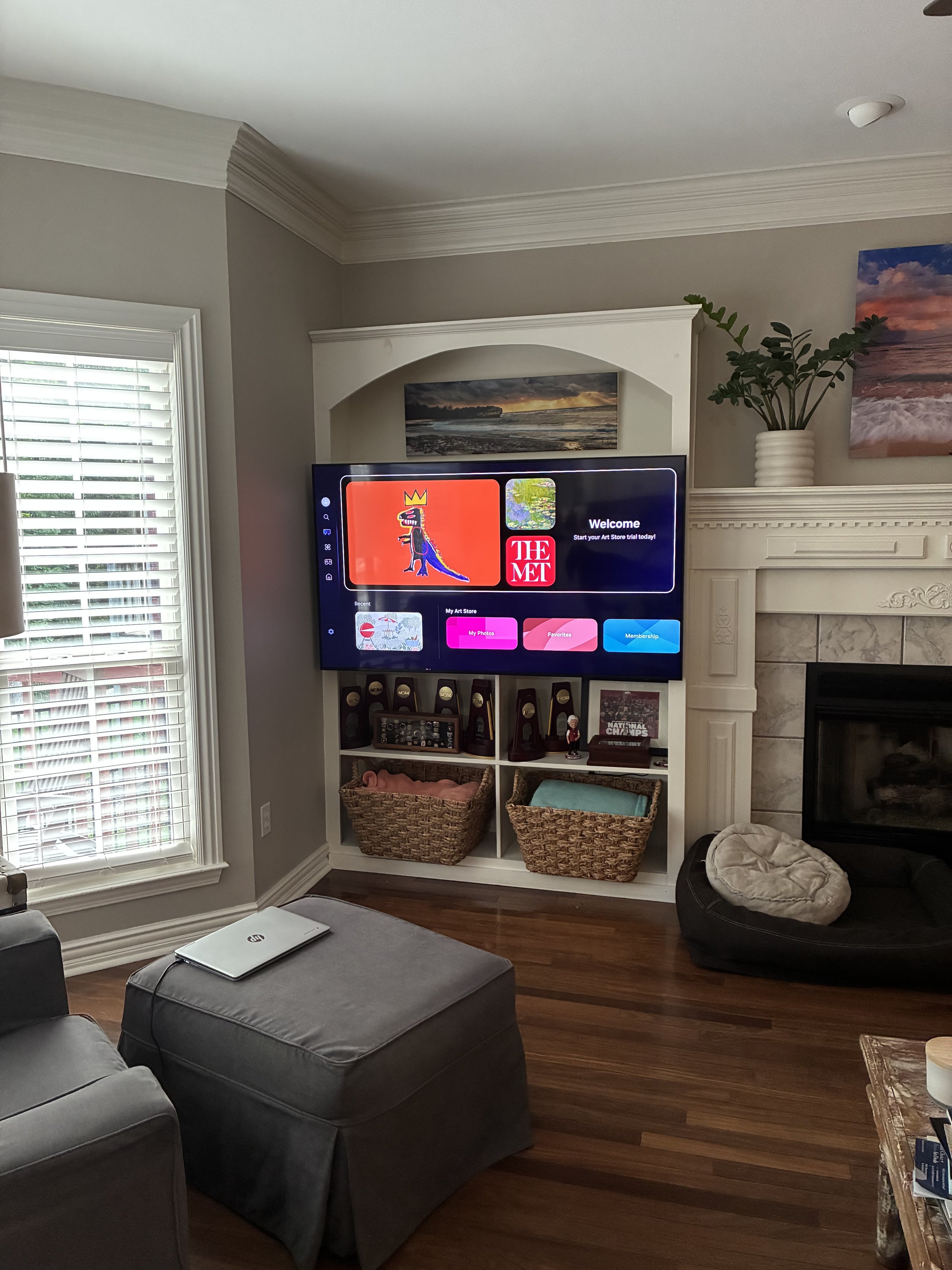 Living room with a wall-mounted TV displaying an art store interface, a window with blinds, a fireplace with a plant on top, and a pet bed on the floor.