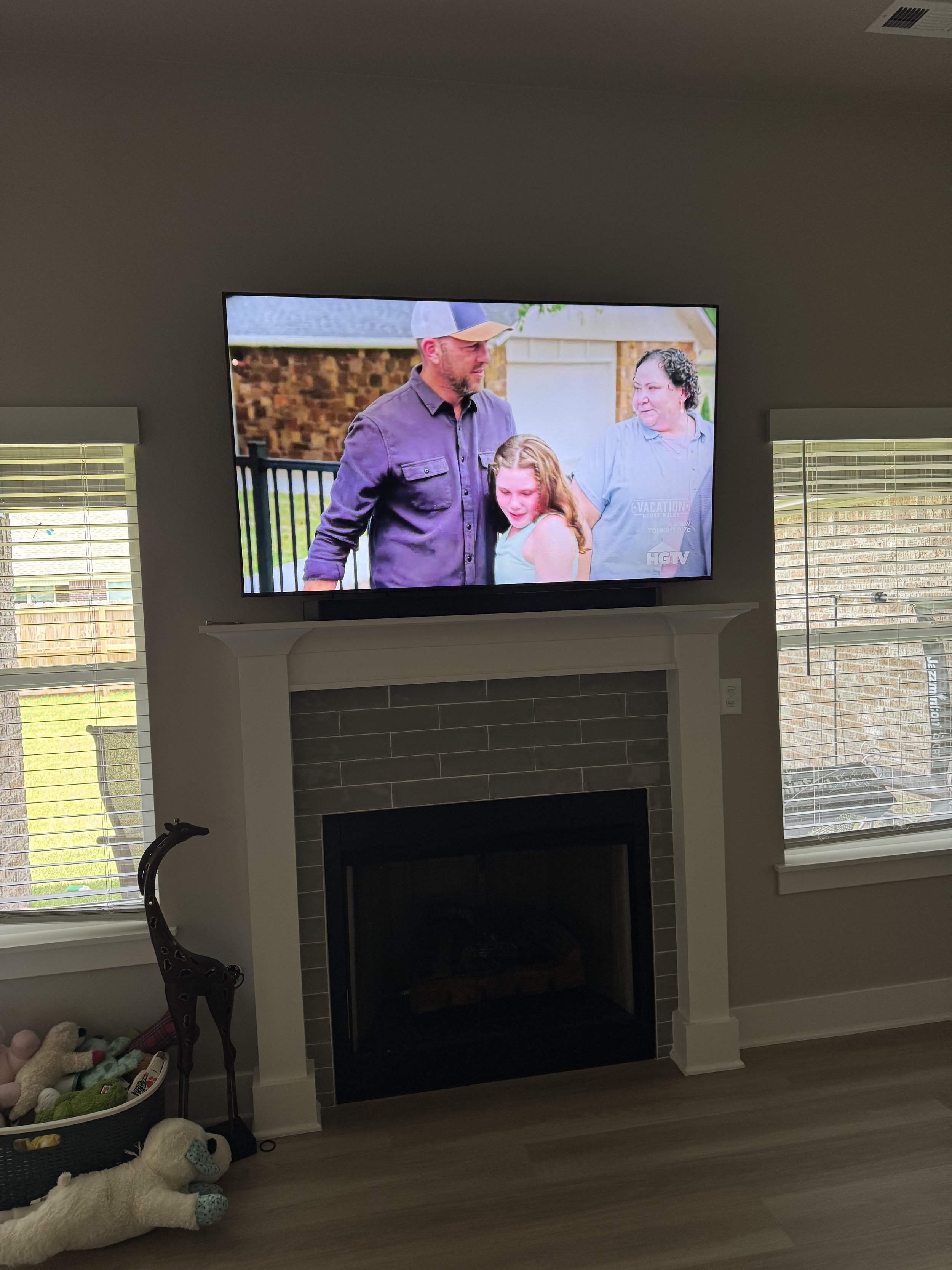 Living room with a wall-mounted flat-screen TV above a fireplace, showing a scene of a man, a woman, and a young girl outdoors. Two windows with blinds on either side of the fireplace, and a basket of stuffed animals and a giraffe figurine on the flo