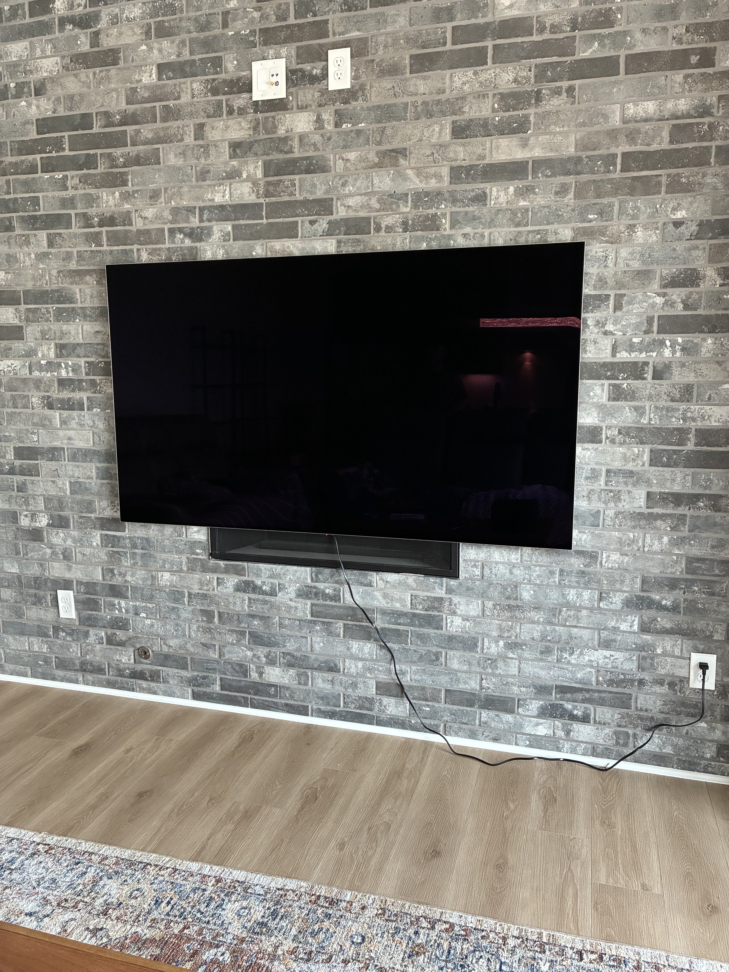 Flat-screen TV mounted on a gray brick wall with outlets and cords visible, situated above a wooden floor and partially over a colorful rug.