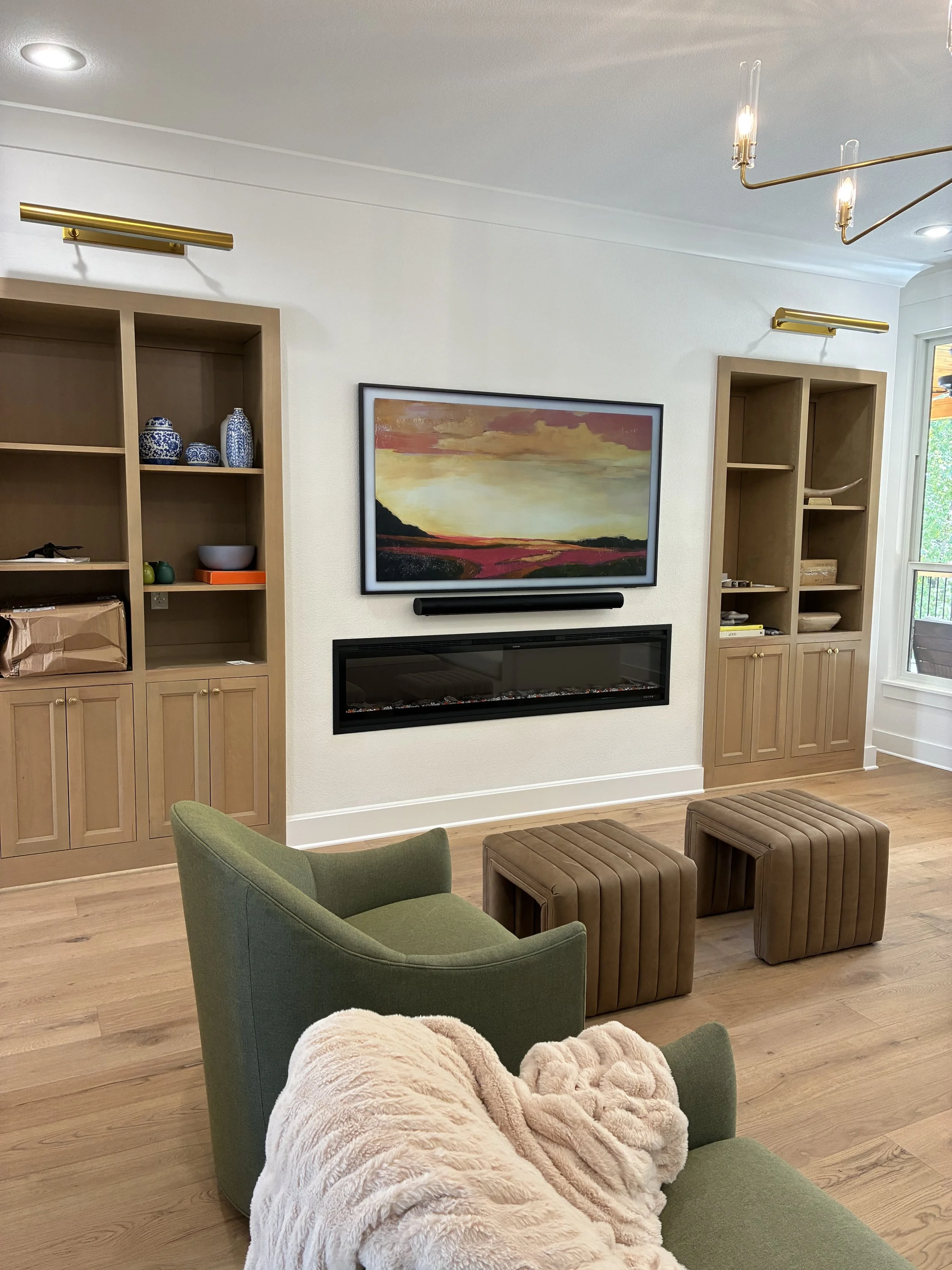 Living room with a framed landscape painting, built-in wooden shelves with decorative items, a green armchair, two brown padded stools, a cream throw blanket, and a modern electric fireplace.