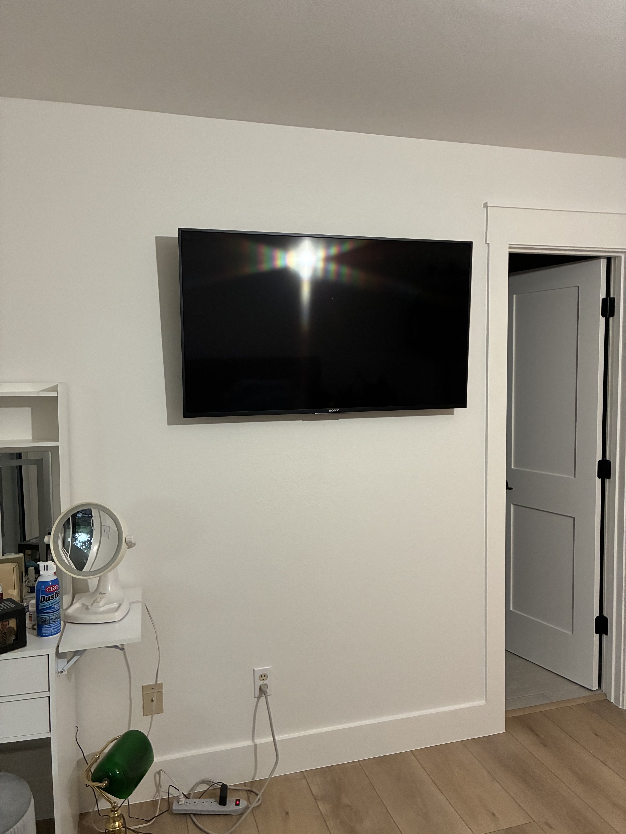 A flat-screen TV mounted on a white wall in a room with a door partially open to the right. A white vanity table with a mirror and various items is visible on the left, with a power strip and cords on the floor.