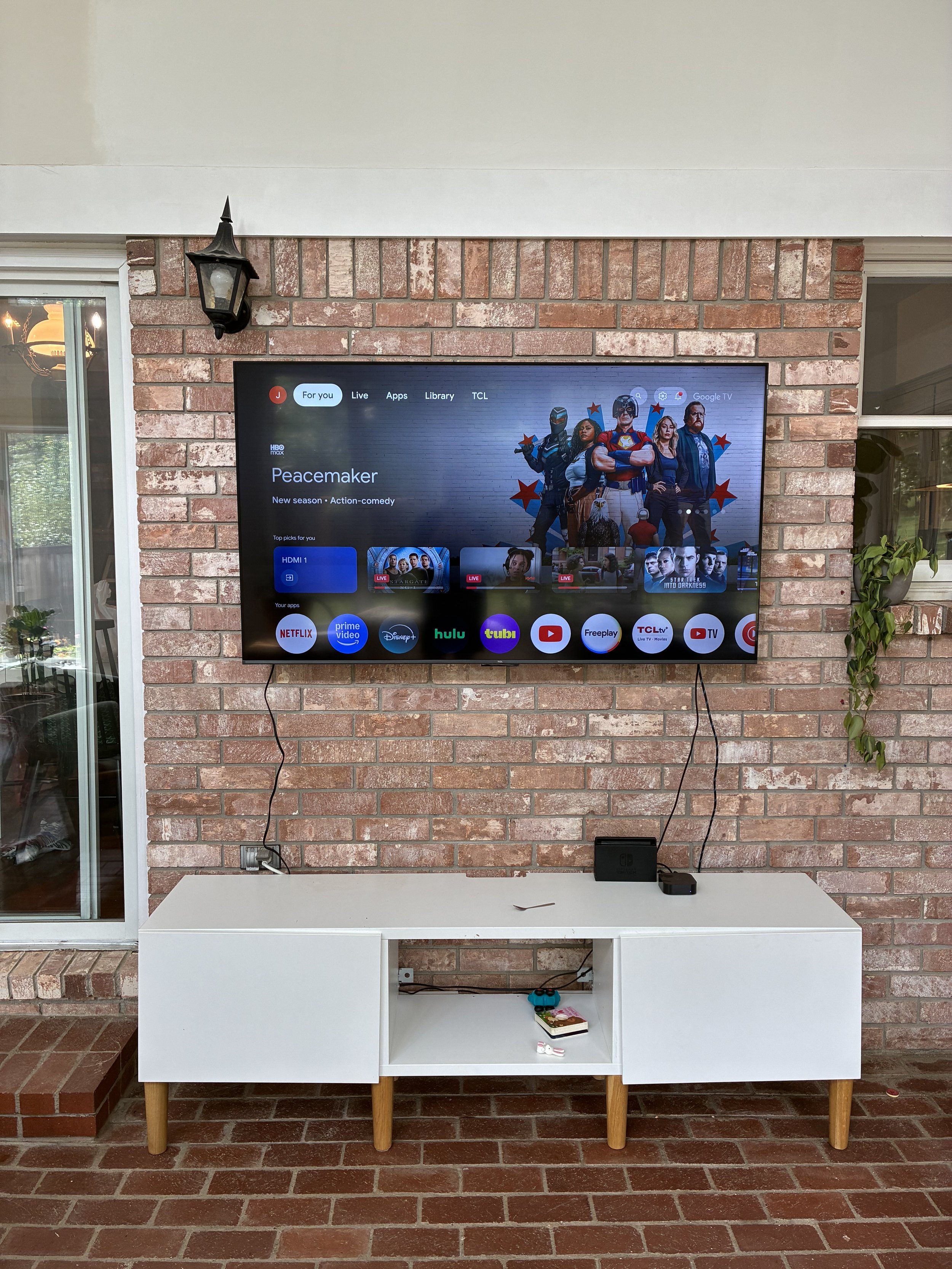 Wall-mounted flat-screen TV displaying the streaming service menu with the show 'Peacemaker' highlighted, mounted on a brick wall with two black cables hanging down, above a white TV stand with a closed compartment, a small device, and some objects o