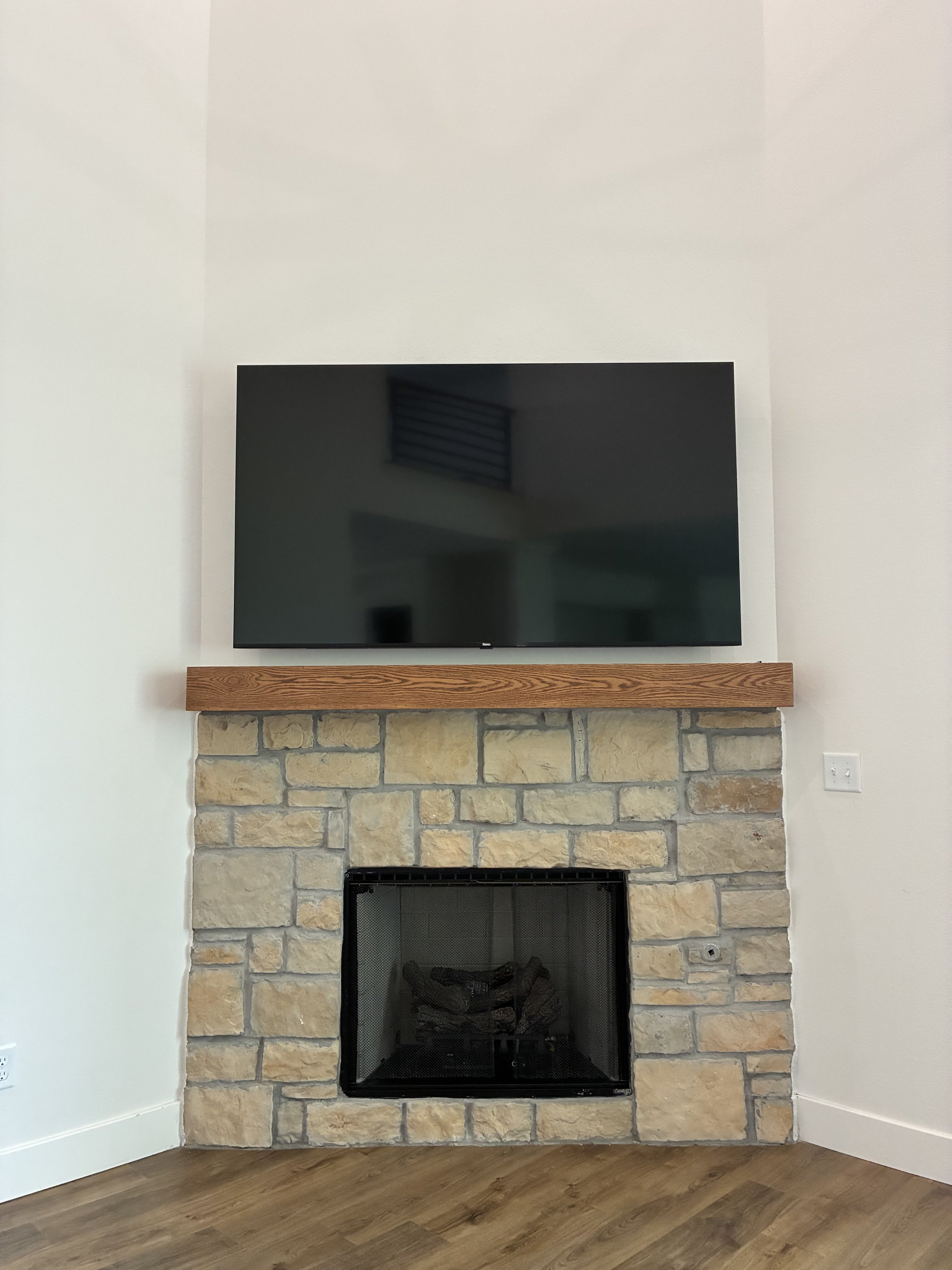 A living room fireplace with a stone surround, a wooden mantle, and a flat-screen TV mounted above it on a white wall.