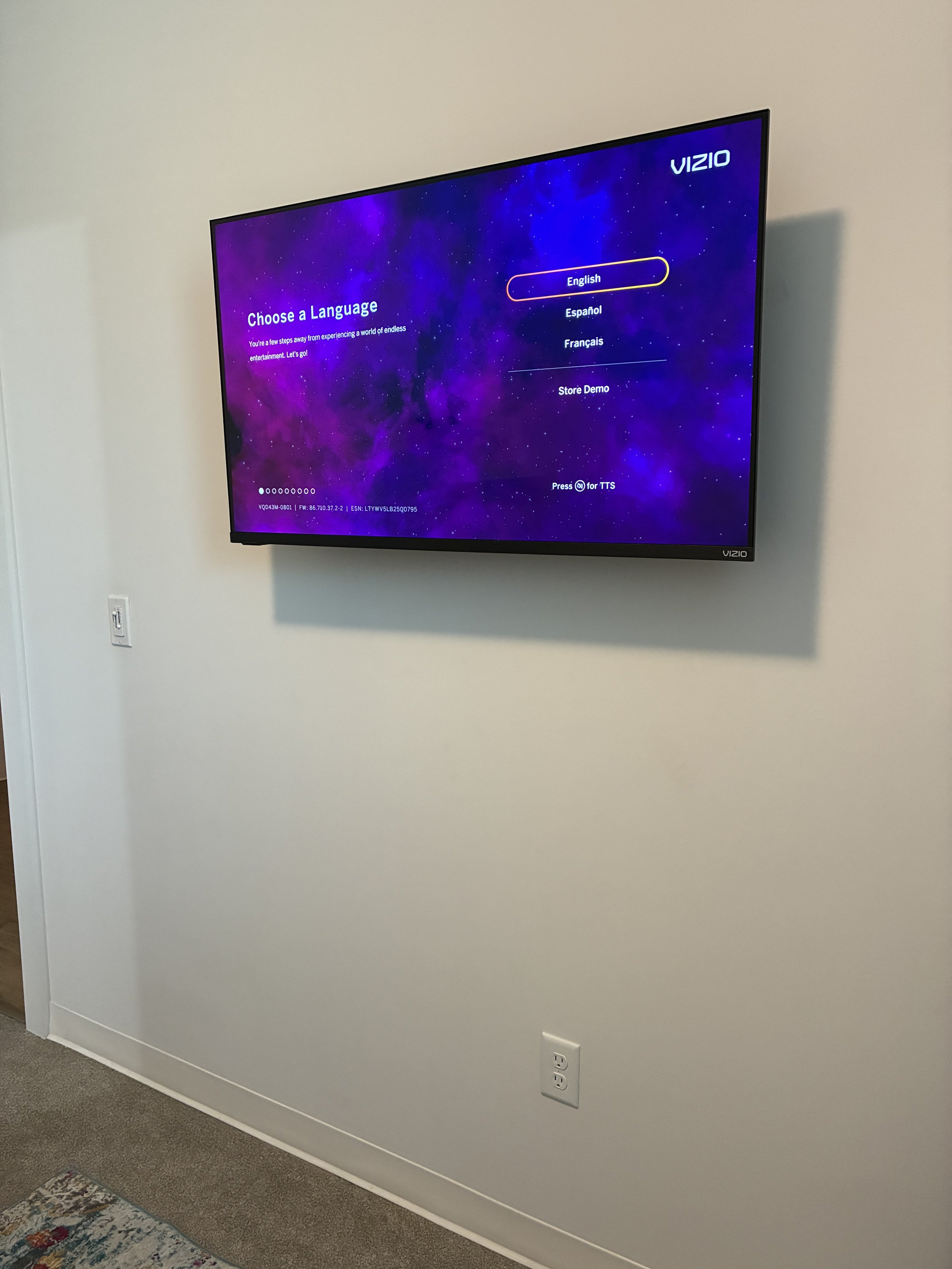 A flat screen TV mounted on a white wall displaying a language selection screen with options for English, Espanol, Francais, and Store Demo, with a purple galaxy-themed background.