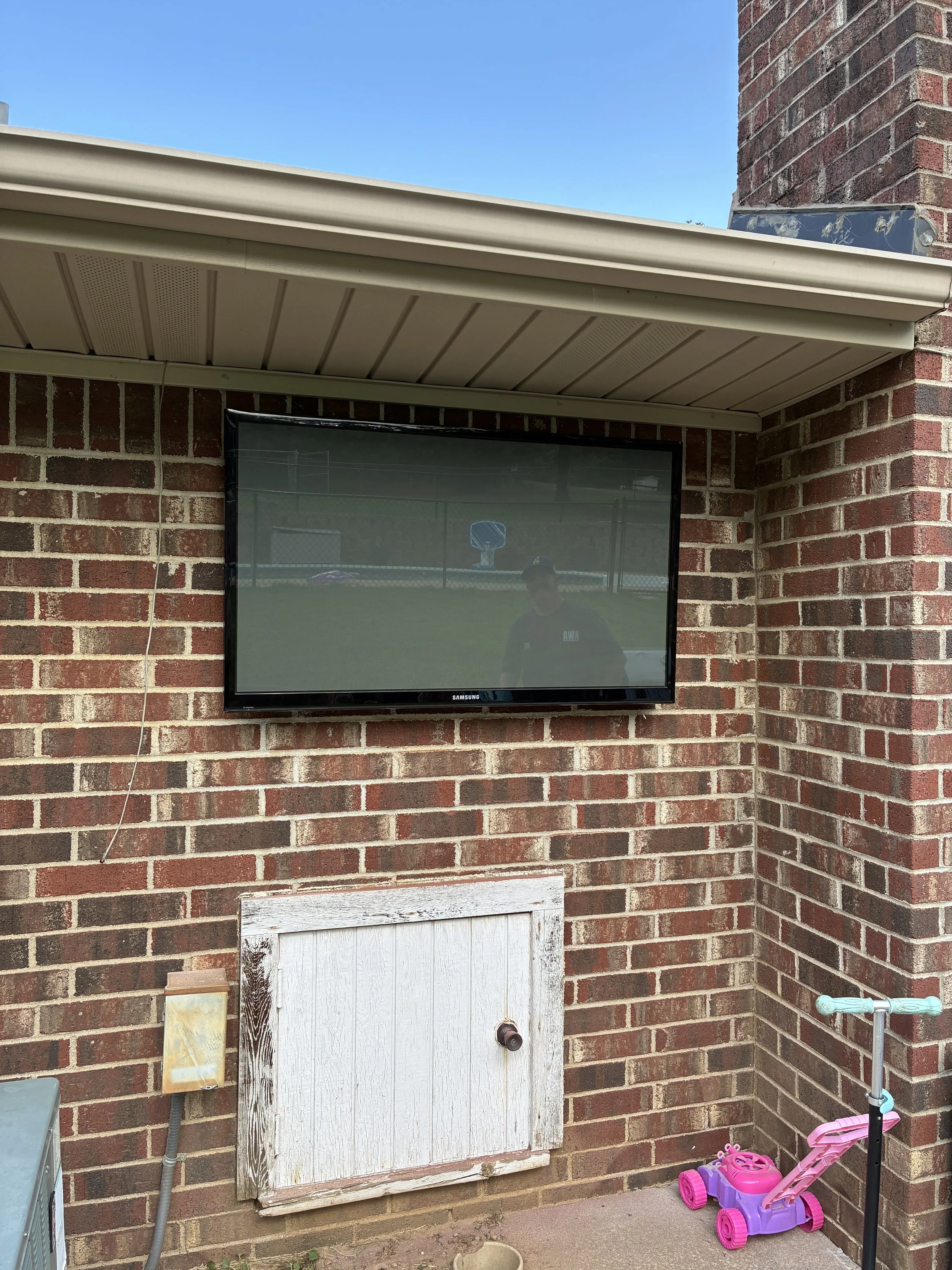 A flat-screen Samsung television mounted on a brick wall outside, with a reflection of a person standing nearby. There is a small door or panel below the TV, a pink and purple children's toy stroller, and a scooter in the corner.