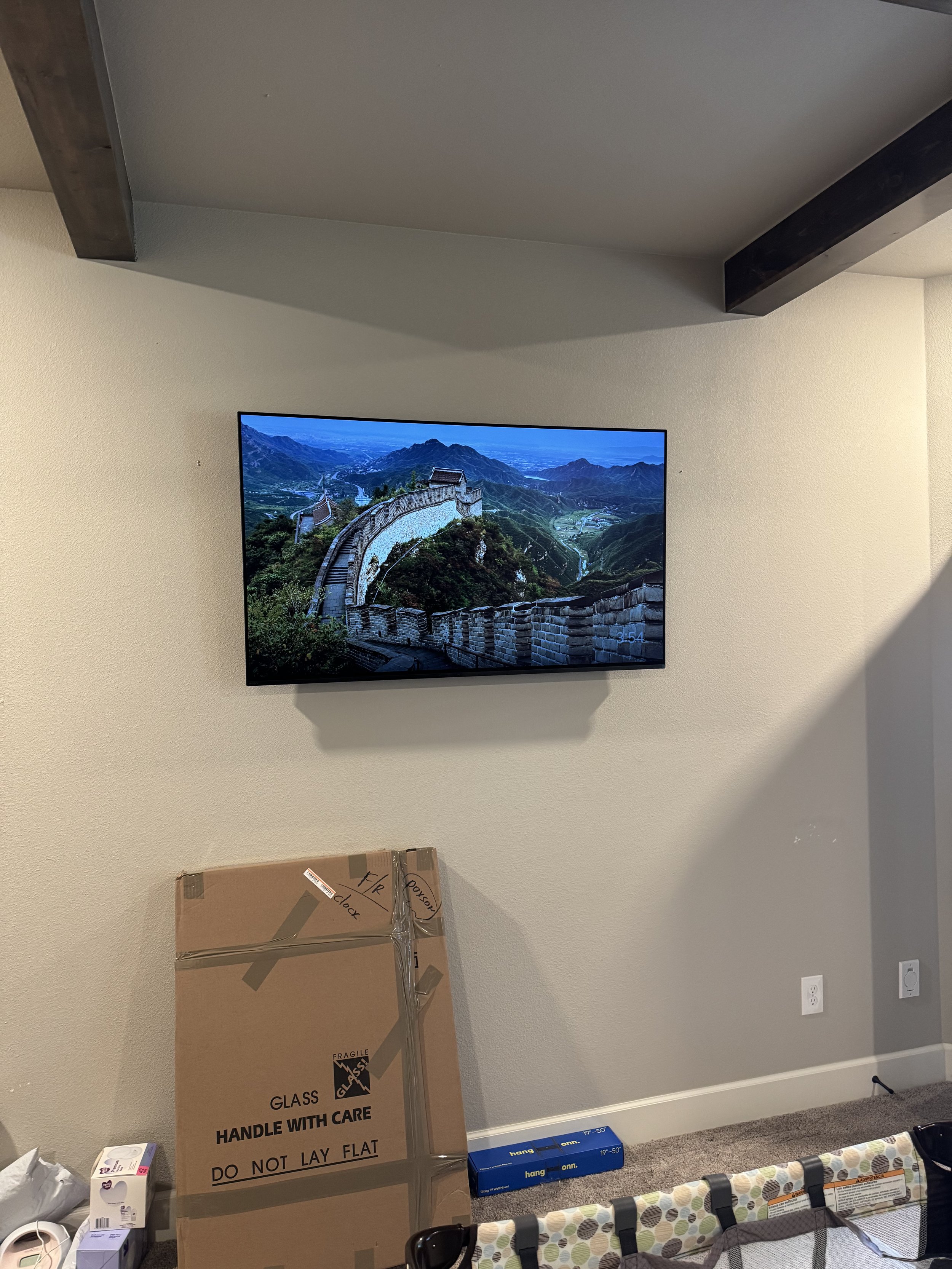 Flat-screen TV mounted on a beige wall displaying an image of the Great Wall of China in a mountainous landscape. Below the TV, there is a large cardboard box labeled "Fragile Glass" and other unpacked items on a carpeted floor.