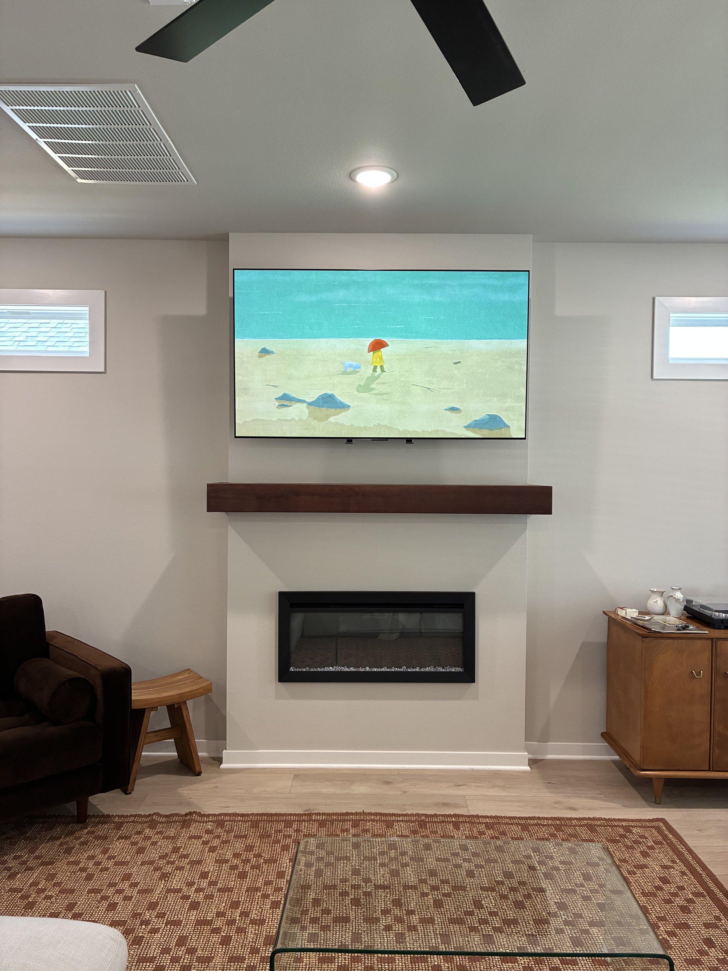 Living room with a wall-mounted TV showing a cartoon beach scene with rocks and a person holding an umbrella, a fireplace below the TV, a brown armchair, a side table, and a wooden cabinet with decorative items.