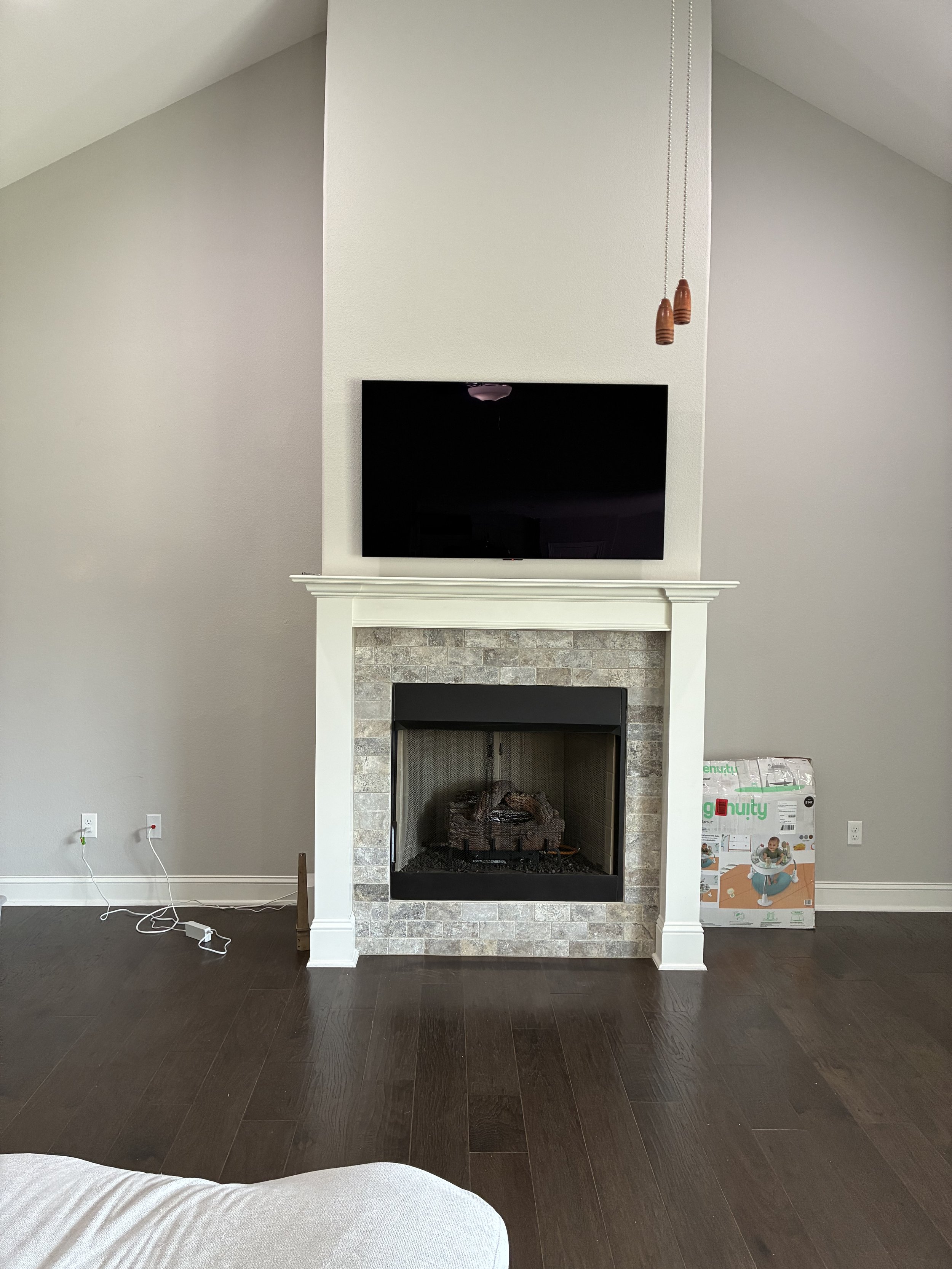 Living room with a fireplace surrounded by stone, a mounted flat-screen TV above it, a box with a child's playpen on the right, and some cords on the left side.