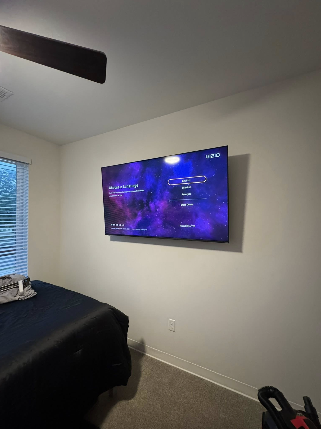 Bedroom with a wall-mounted Vizio smart TV displaying a language selection screen against a plain beige wall, a bed with a black bedspread and a gray bag, a window with blinds, and a ceiling fan.
