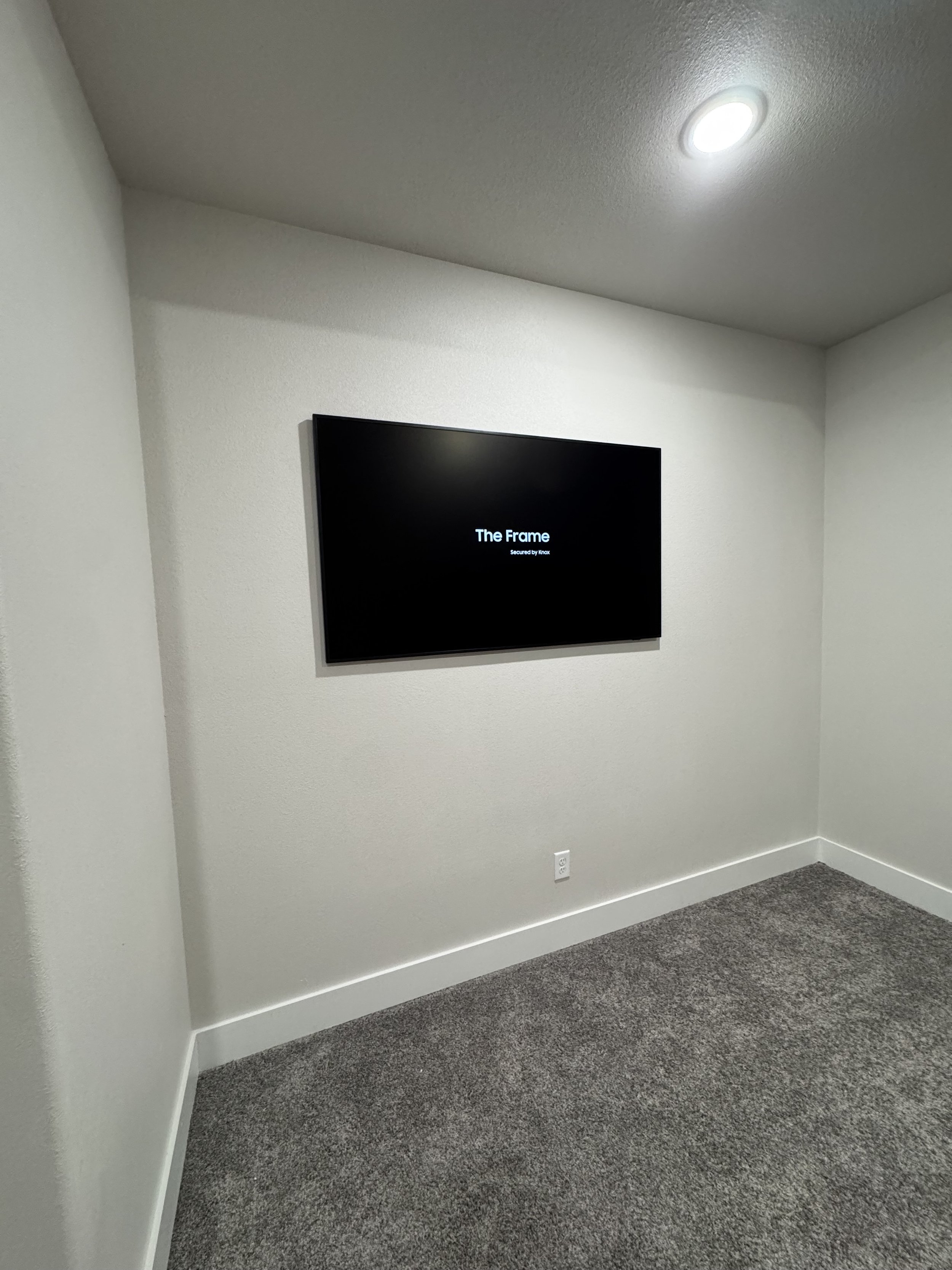 Empty room with a wall-mounted TV displaying 'The Frame Secured by Knox' on a black screen, white walls, gray carpet, and a ceiling light.