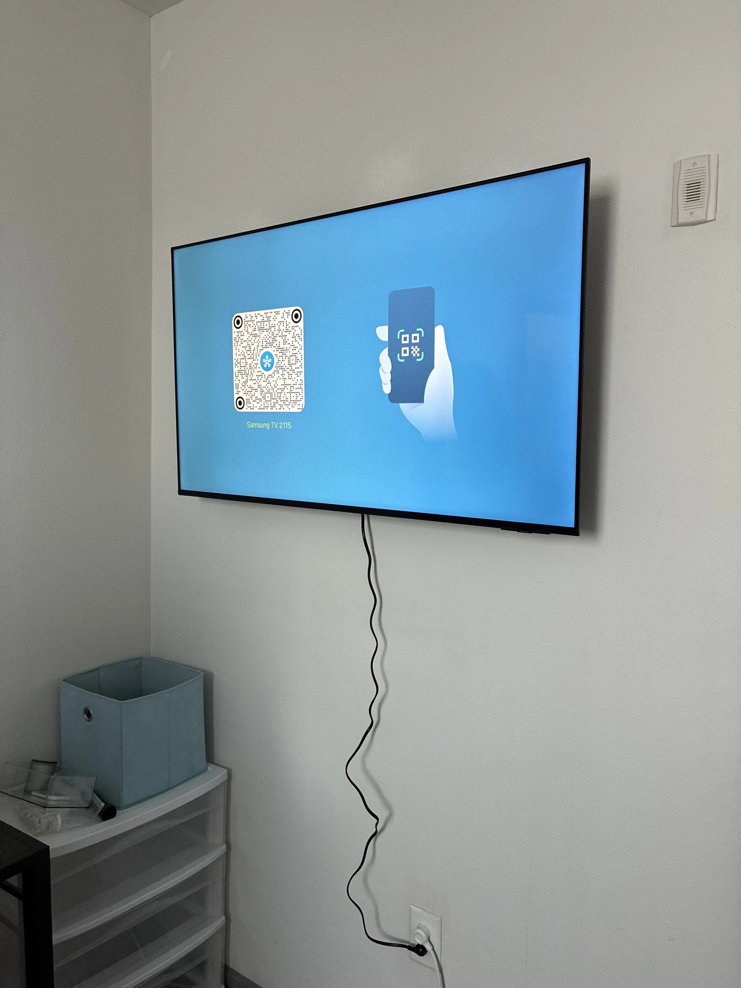 A TV mounted on a white wall displaying a QR code and an illustration of a hand holding a smartphone. A black cable hangs from the TV, plugged into an outlet. There is a blue storage box on a white plastic drawer unit below the TV.