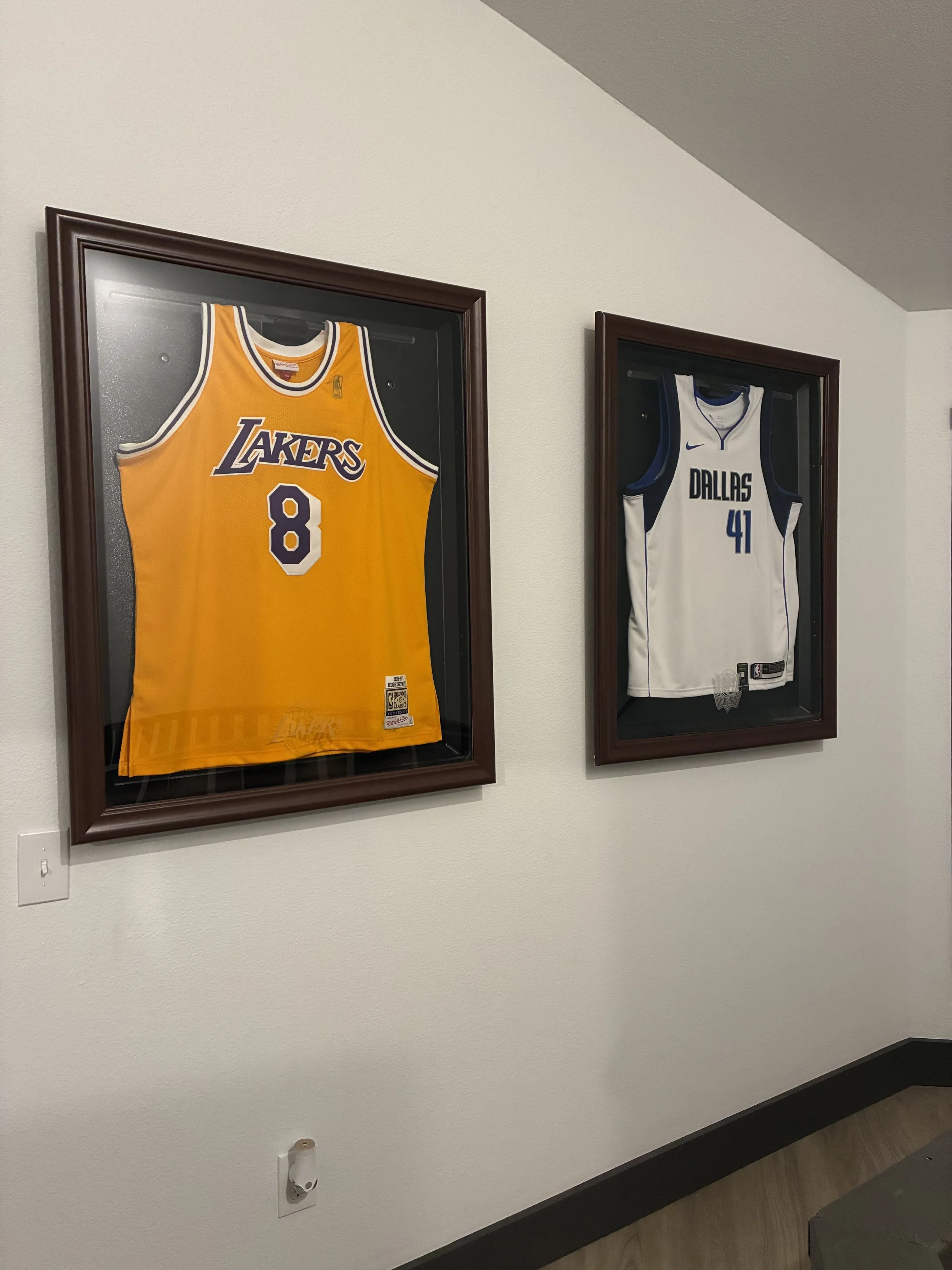 Two framed basketball jerseys hanging on a wall, one from the Los Angeles Lakers with number 8, yellow and purple colors, and another from the Dallas Mavericks with number 41, white with blue accents.