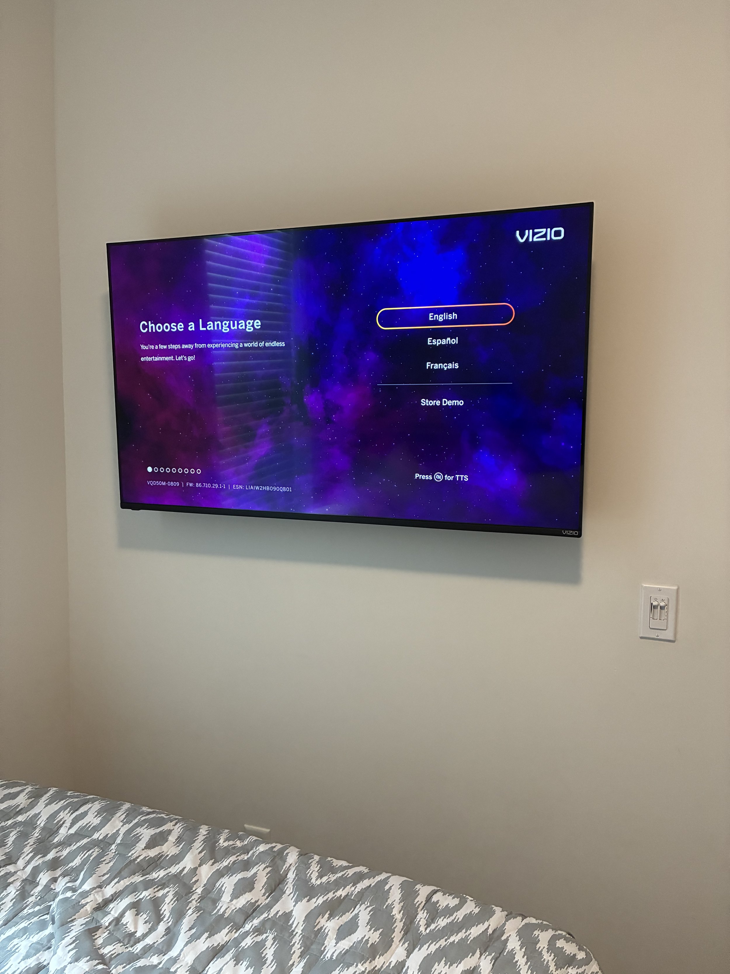 Flat-screen TV mounted on a white wall displaying a language selection screen with options: English, Español, Français, and Store Demo, with the Vizio logo in the top right corner. Part of a bed with a patterned gray and white bedspread is visible in