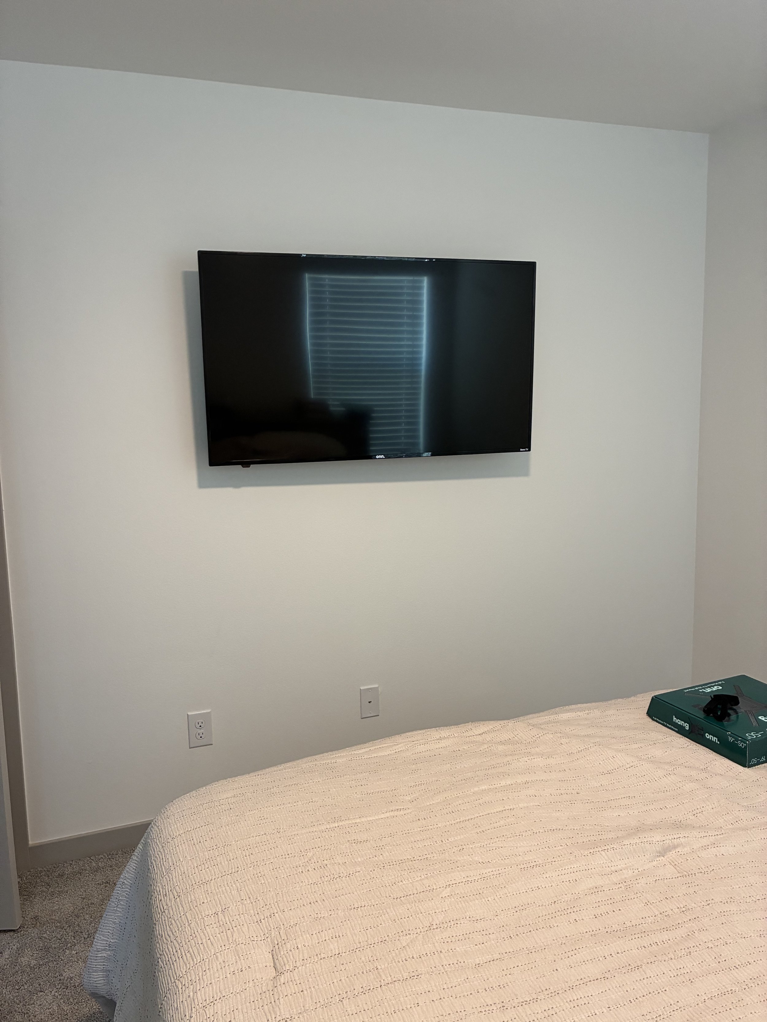 Bedroom with flat-screen TV mounted on the white wall, beige bedspread on the bed, and a box on the bed.