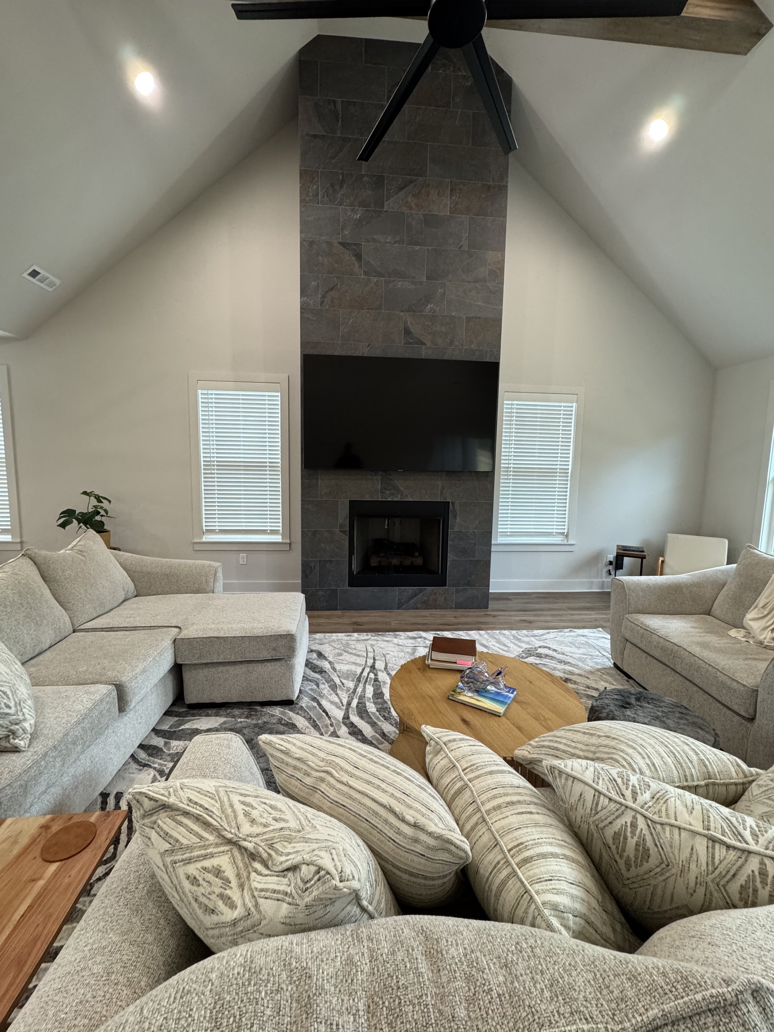 Living room with a high ceiling, fireplace with a TV mounted above, white walls, two windows with blinds, a beige sectional sofa with multiple pillows, a round wooden coffee table with books, and a gray and white patterned rug.