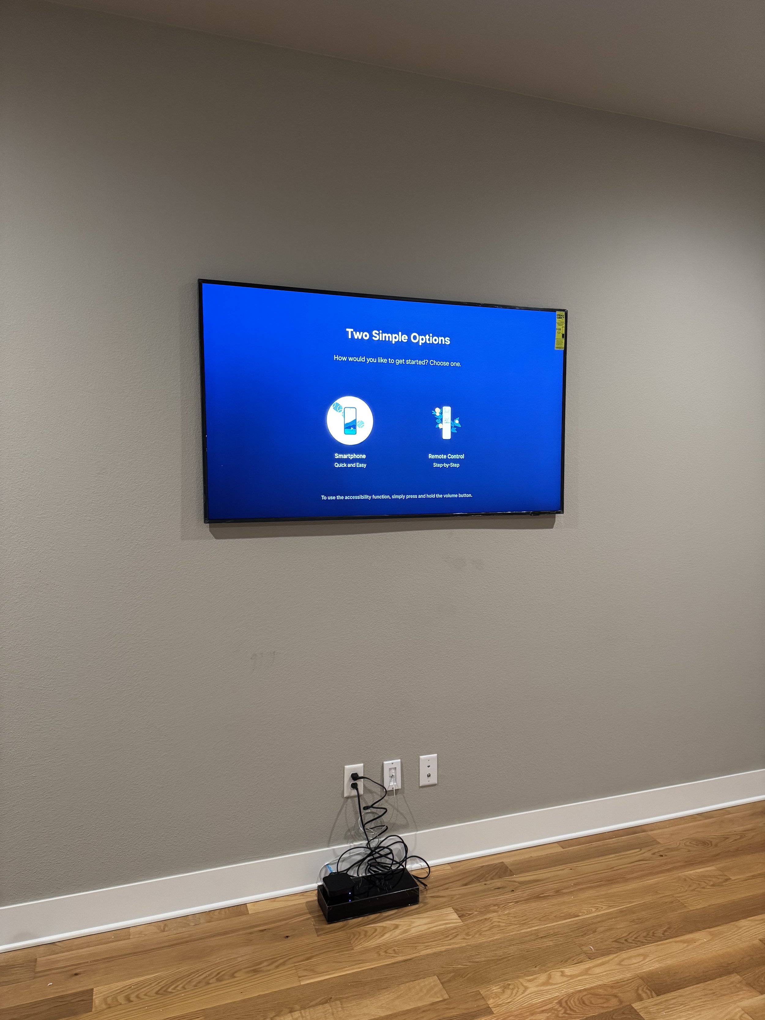 Wall-mounted flat-screen television displaying setup options, connected to a small black device with tangled cables, on a wooden floor in a room with plain gray walls.