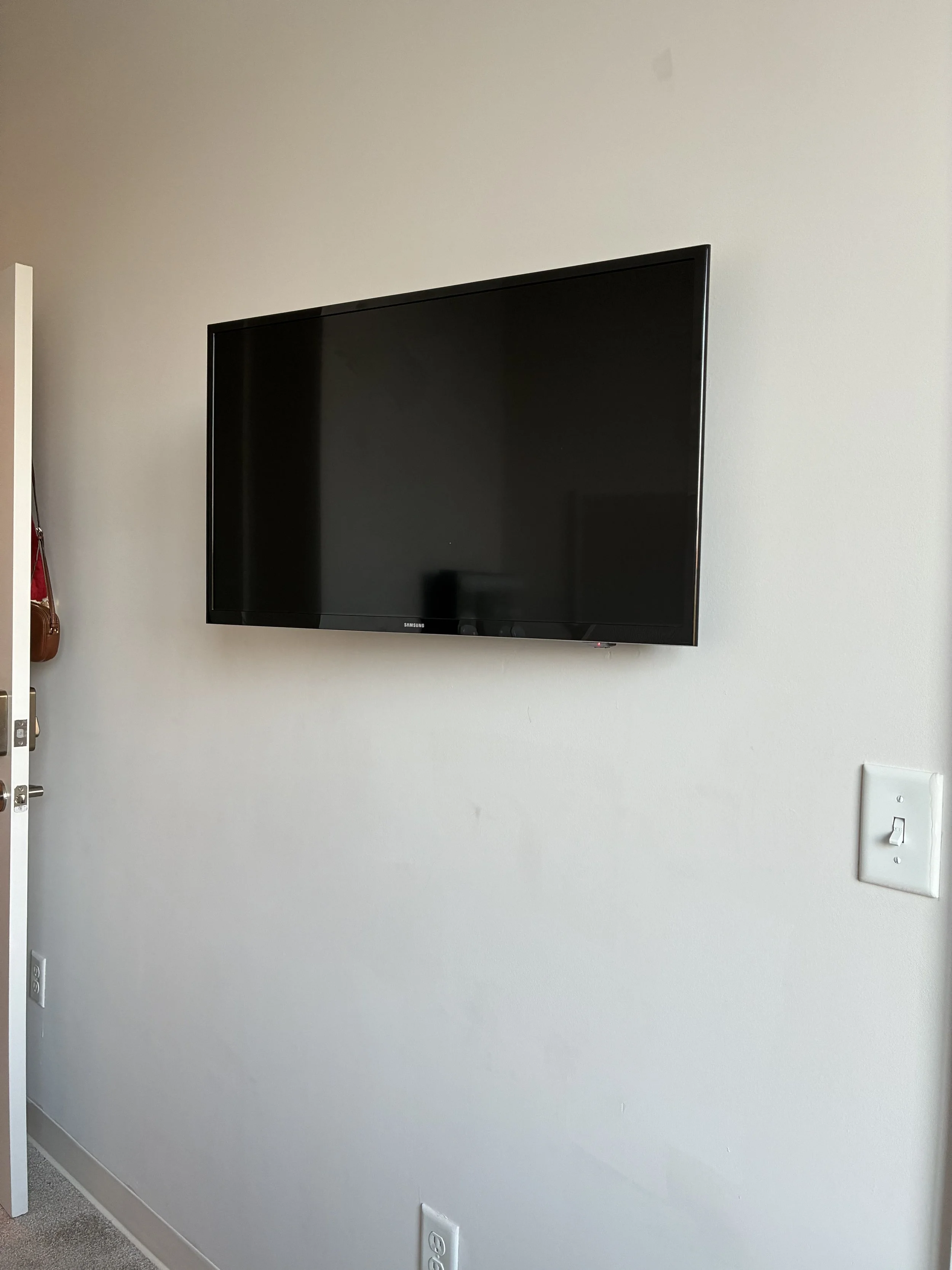 Flat-screen television mounted on a white wall in a room, with a light switch and electrical outlet visible nearby.