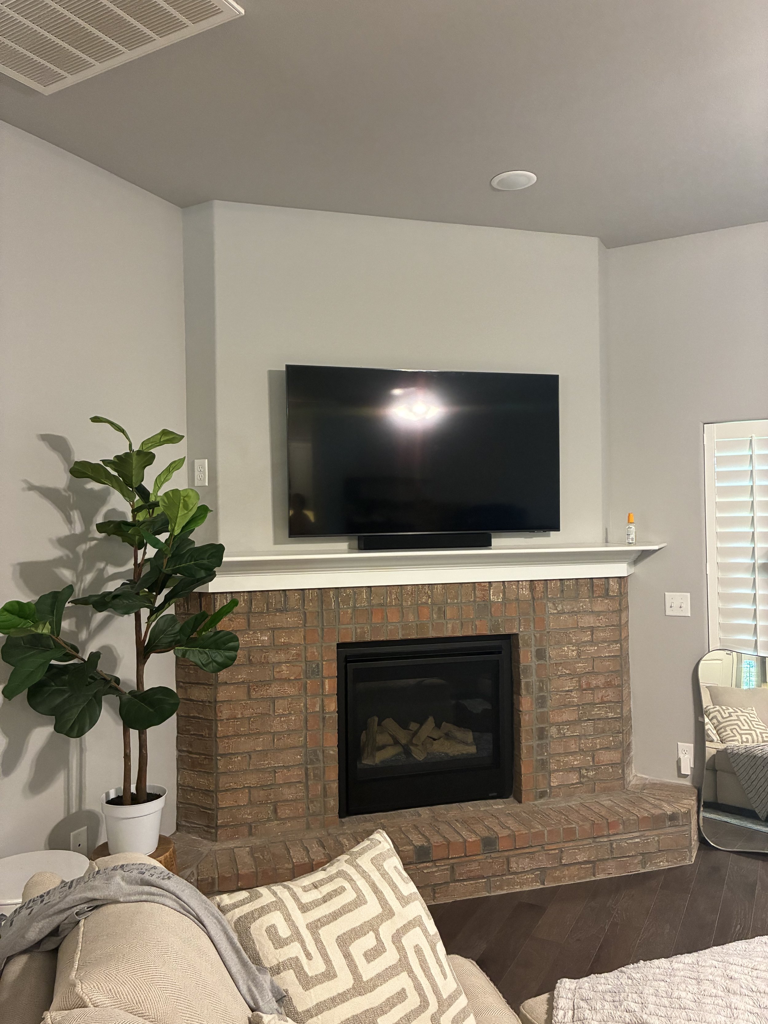 Living room with a brick fireplace, a wall-mounted flat-screen TV above it, a large green potted plant next to the fireplace, a beige couch with patterned pillows, a mirror, and a window with shutters.
