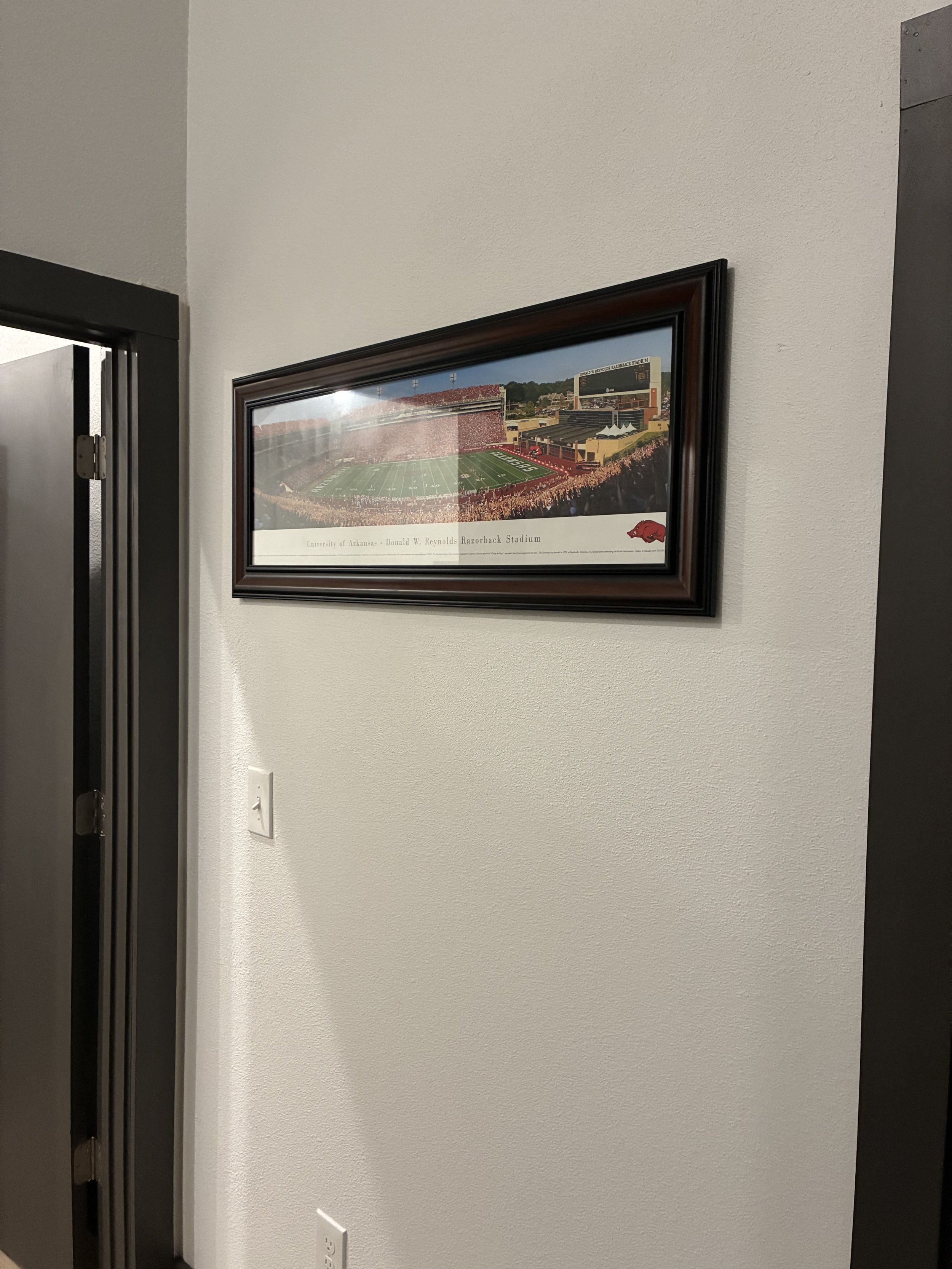 Photograph of a framed poster on a white wall, showing a stadium with a football field and crowd, labeled as Razorback Stadium at the University of Arkansas.