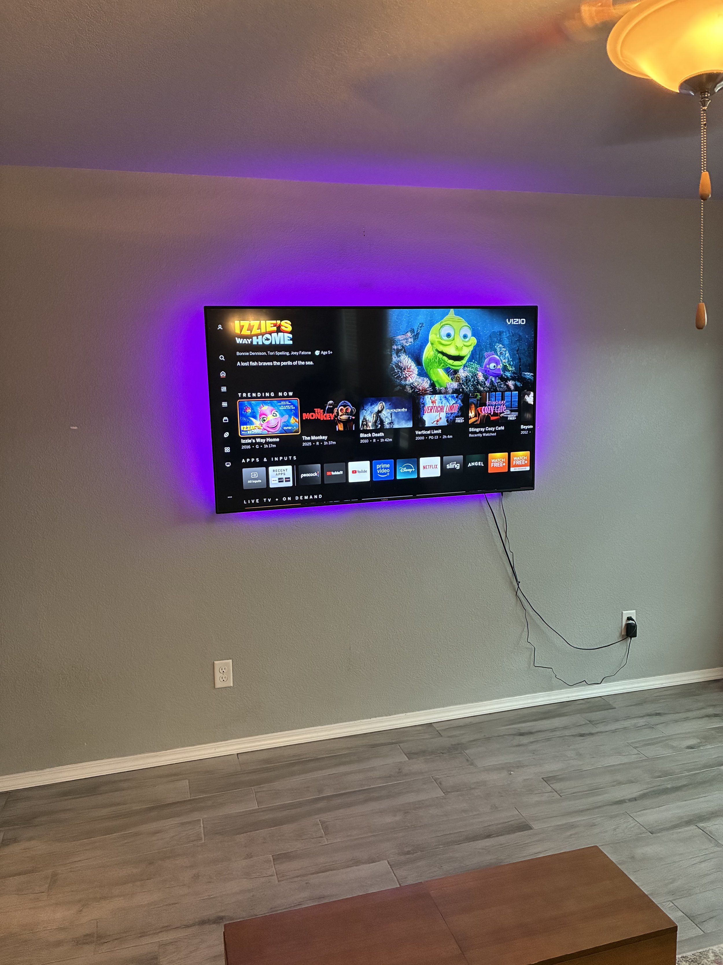 Wall-mounted flat-screen TV displaying a streaming service menu with a cartoon character on the screen, purple ambient lighting behind the TV, a ceiling fan with a yellowish light, electrical outlet and power cord visible, wood and laminate flooring,