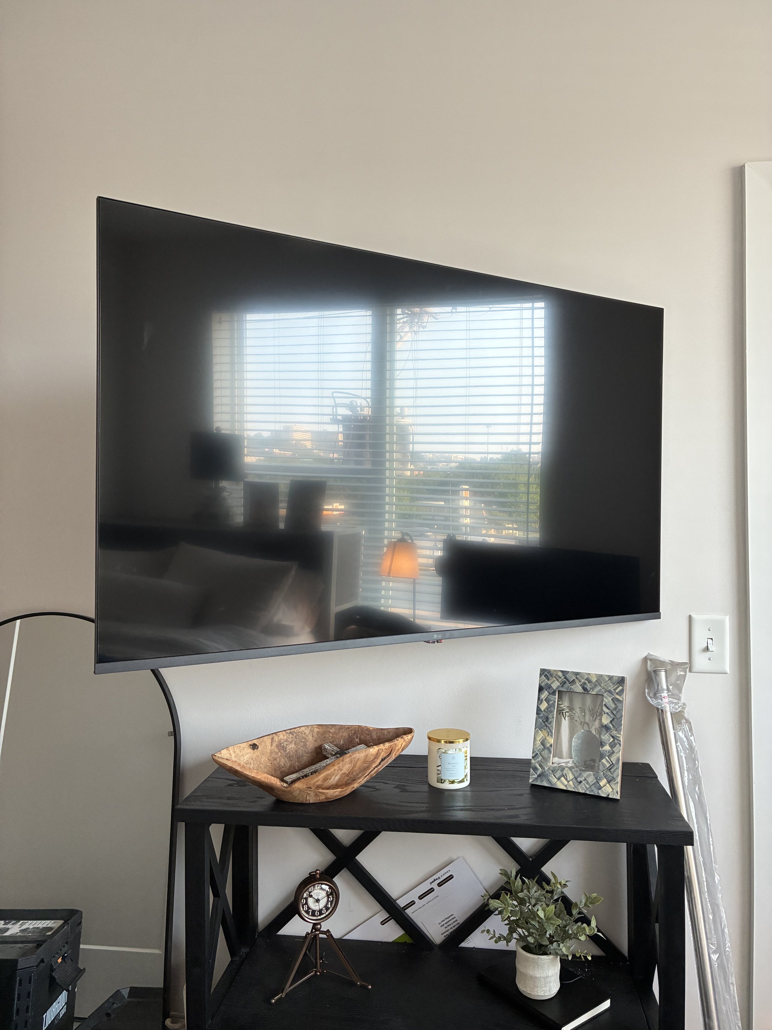 Living room with a large wall-mounted TV reflecting a window with blinds, a wooden bowl, a candle, a picture frame, a plant, and a clock on a black table.