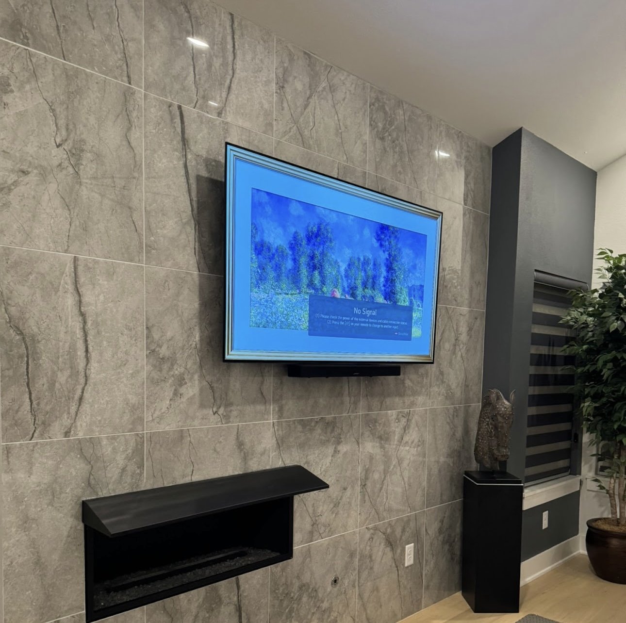 A television on a gray marble wall displaying a 'No Signal' message, with a black shelf below and a decorative sculpture on a black pedestal beside a potted plant.