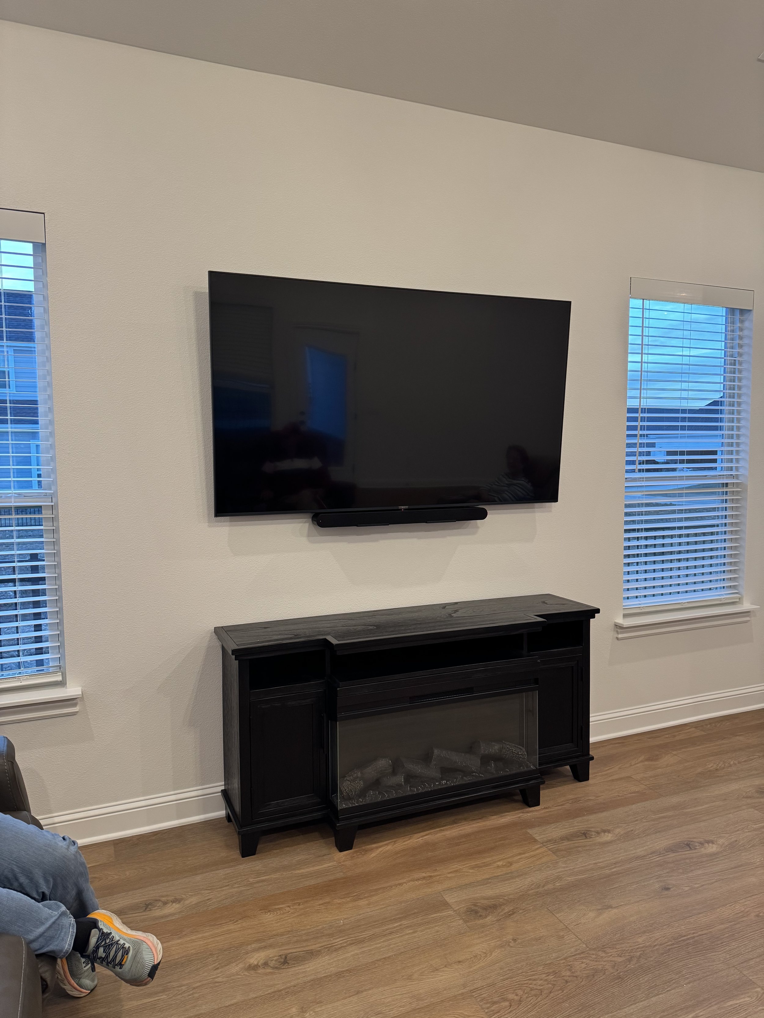 Living room with wall-mounted flat-screen TV, black fireplace below, two windows with blinds, and part of a person's leg and sneaker visible on the left.