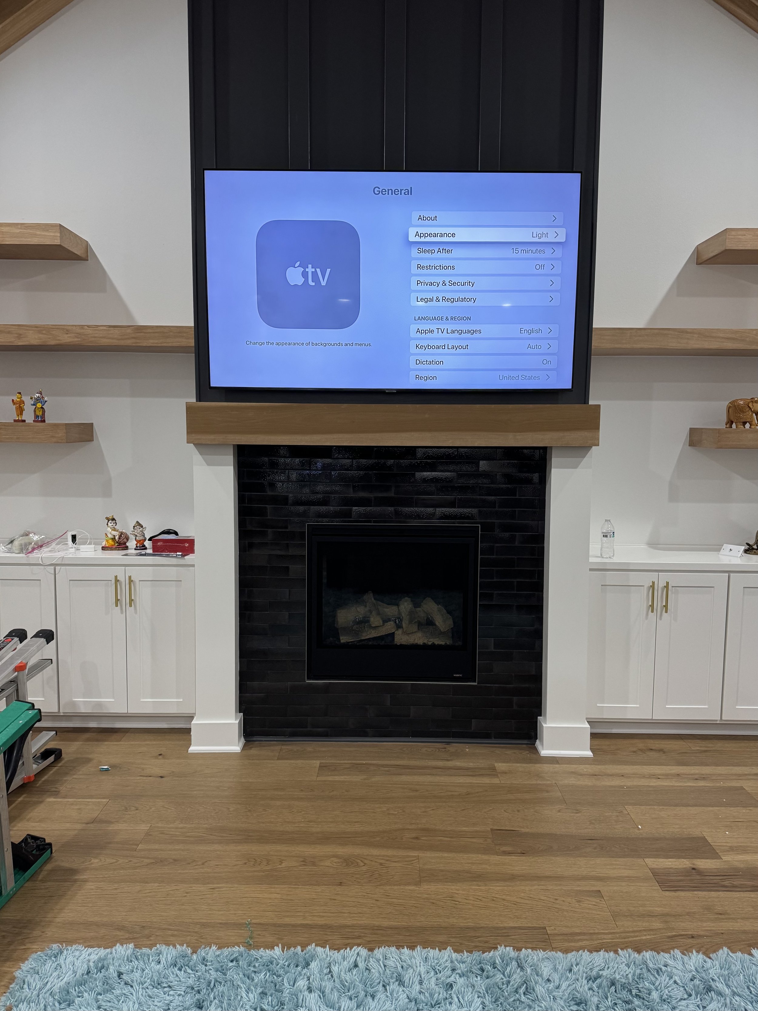 A large flat-screen TV mounted above a fireplace with black brick surround, showing TV settings menu, in a modern living room with white cabinets, wooden shelves, and a blue rug.