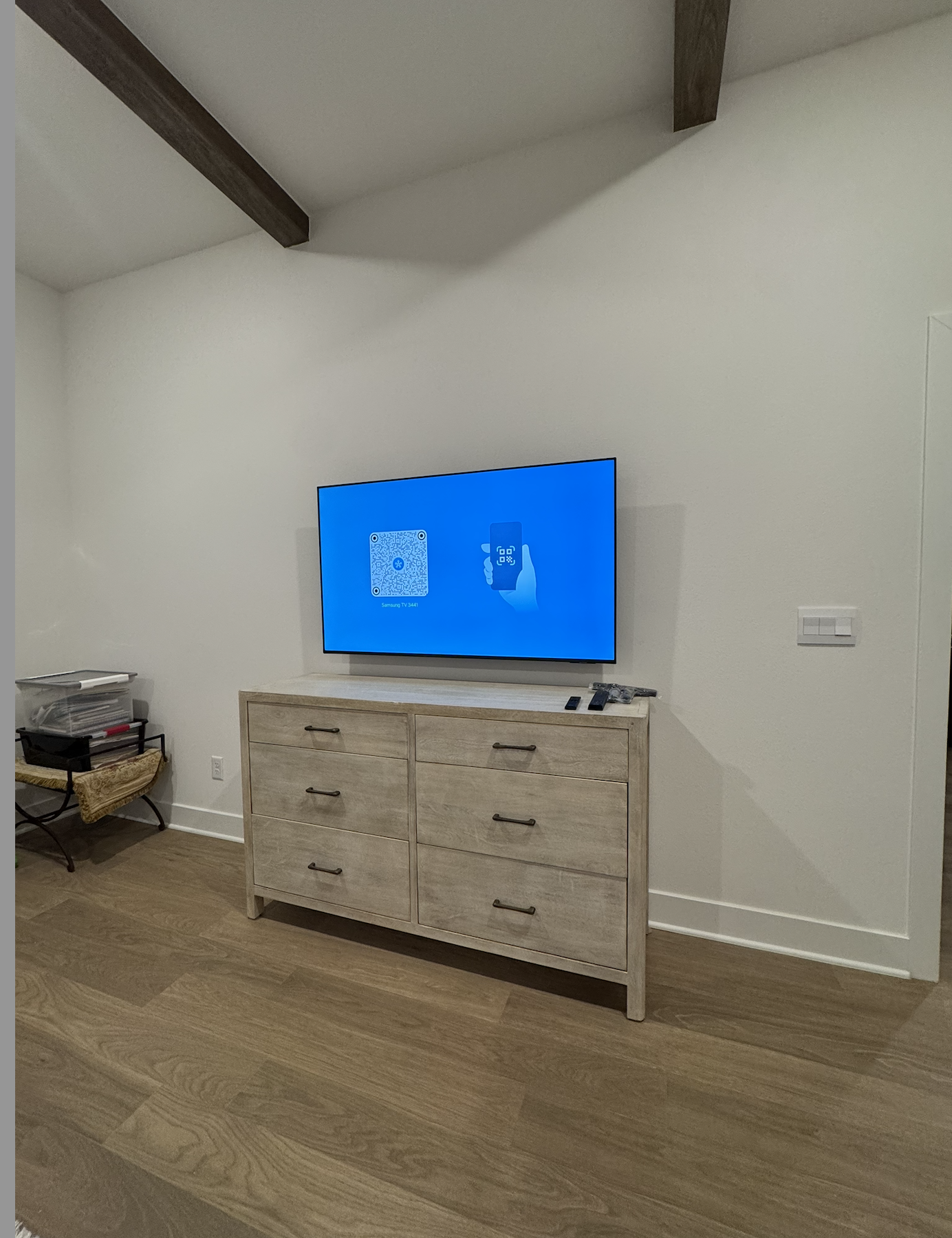 Living room with a mounted flat-screen TV displaying a QR code, a wooden dresser underneath with remote controls, and a small black and glass side table with storage containers on the left side of the room.