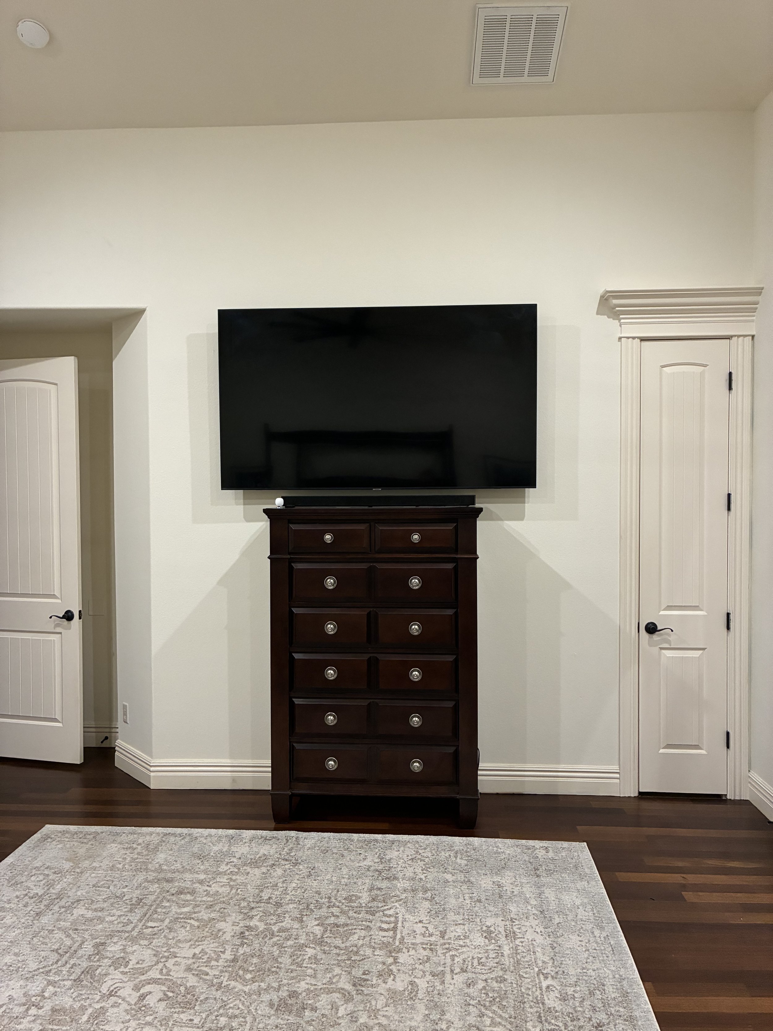 A room with a mounted flat-screen TV above a dark wooden dresser with multiple drawers. The room features white walls, white trim, two white paneled doors, wooden flooring, and a light-colored area rug.