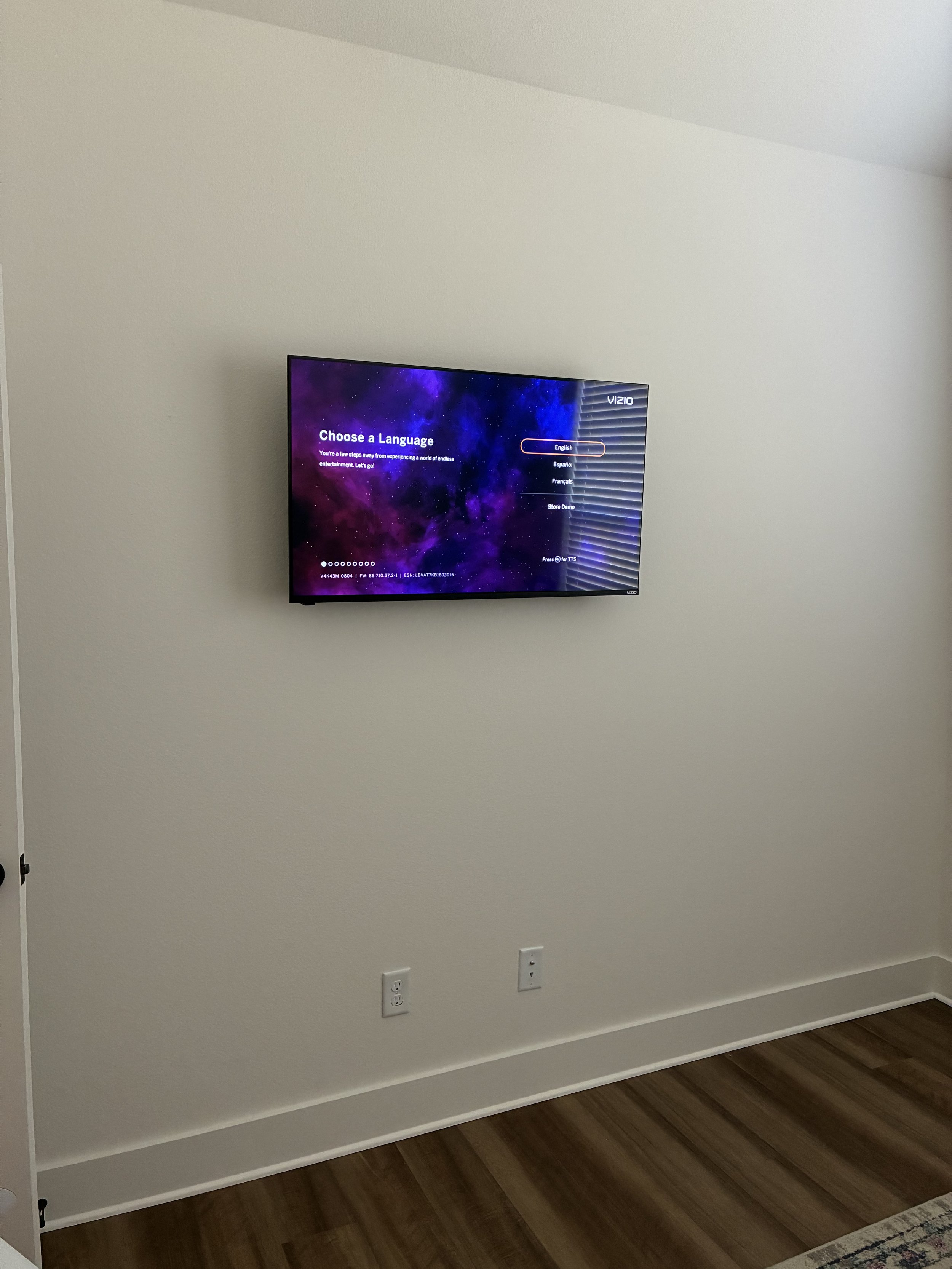 A flat-screen television mounted on a beige wall displaying a language selection menu. The room has wood flooring and two electrical outlets below the TV.