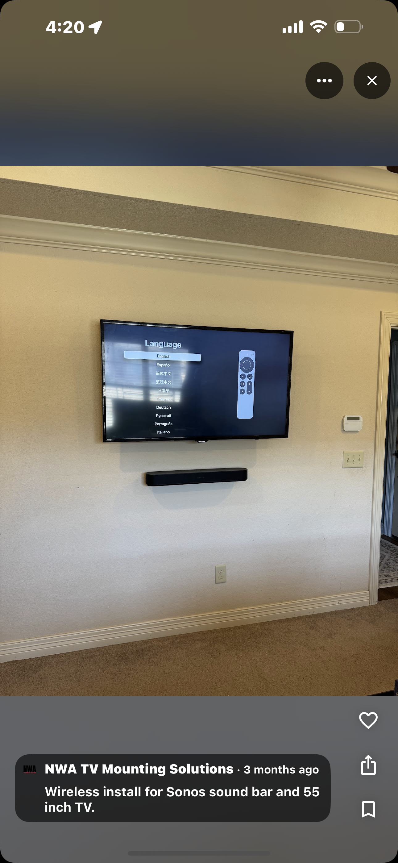 A flat-screen TV mounted on a beige wall displaying a language selection menu with options including English and Spanish. A soundbar is mounted directly below the TV, and the room features beige carpet and crown molding. A thermostat and a light swit