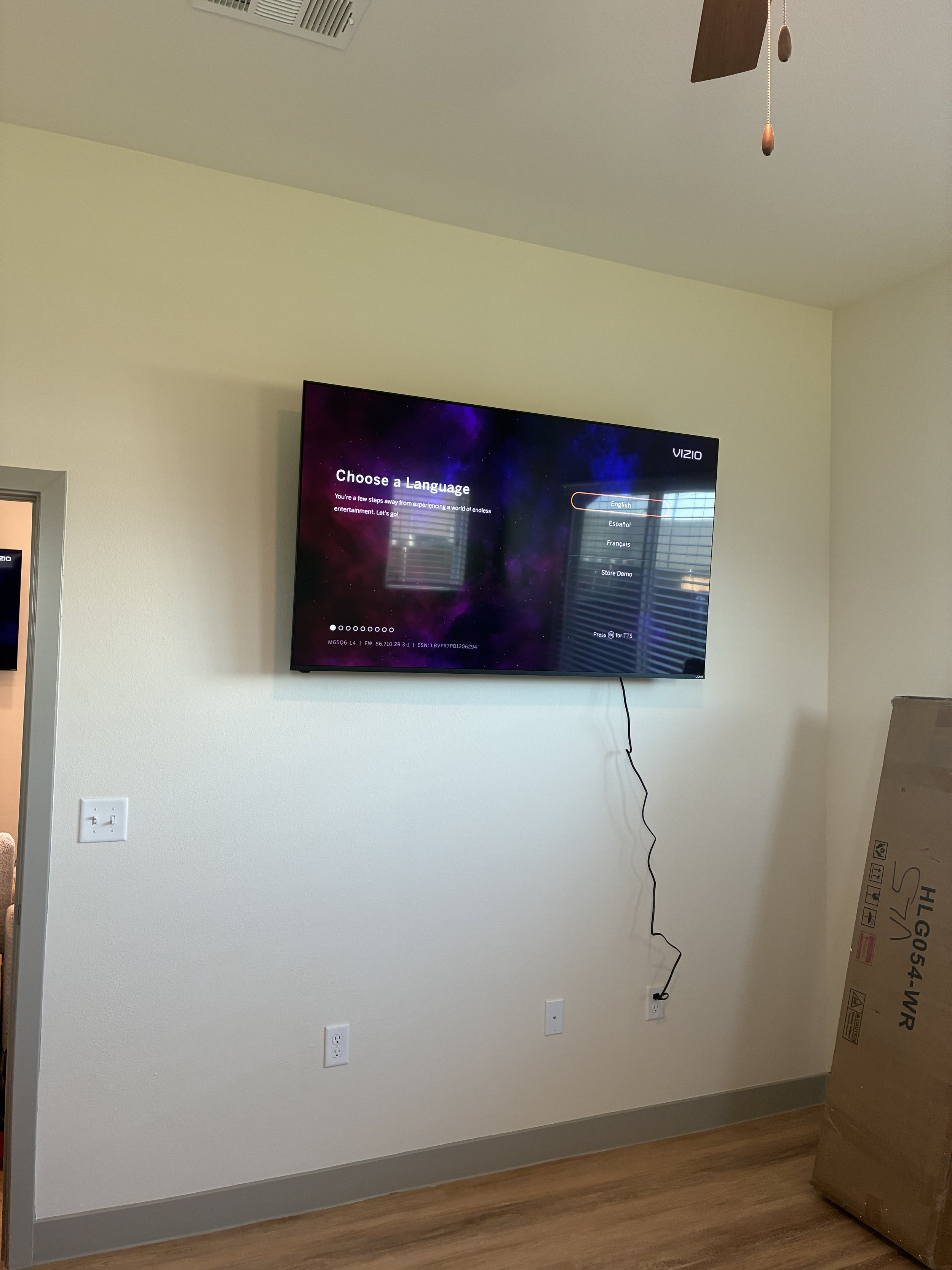 A wall-mounted Vizio television displaying a 'Choose a Language' setup screen in a room with beige walls, light brown flooring, and a ceiling fan. There are multiple electrical outlets and a loose cable hanging from the TV to the outlet. To the right