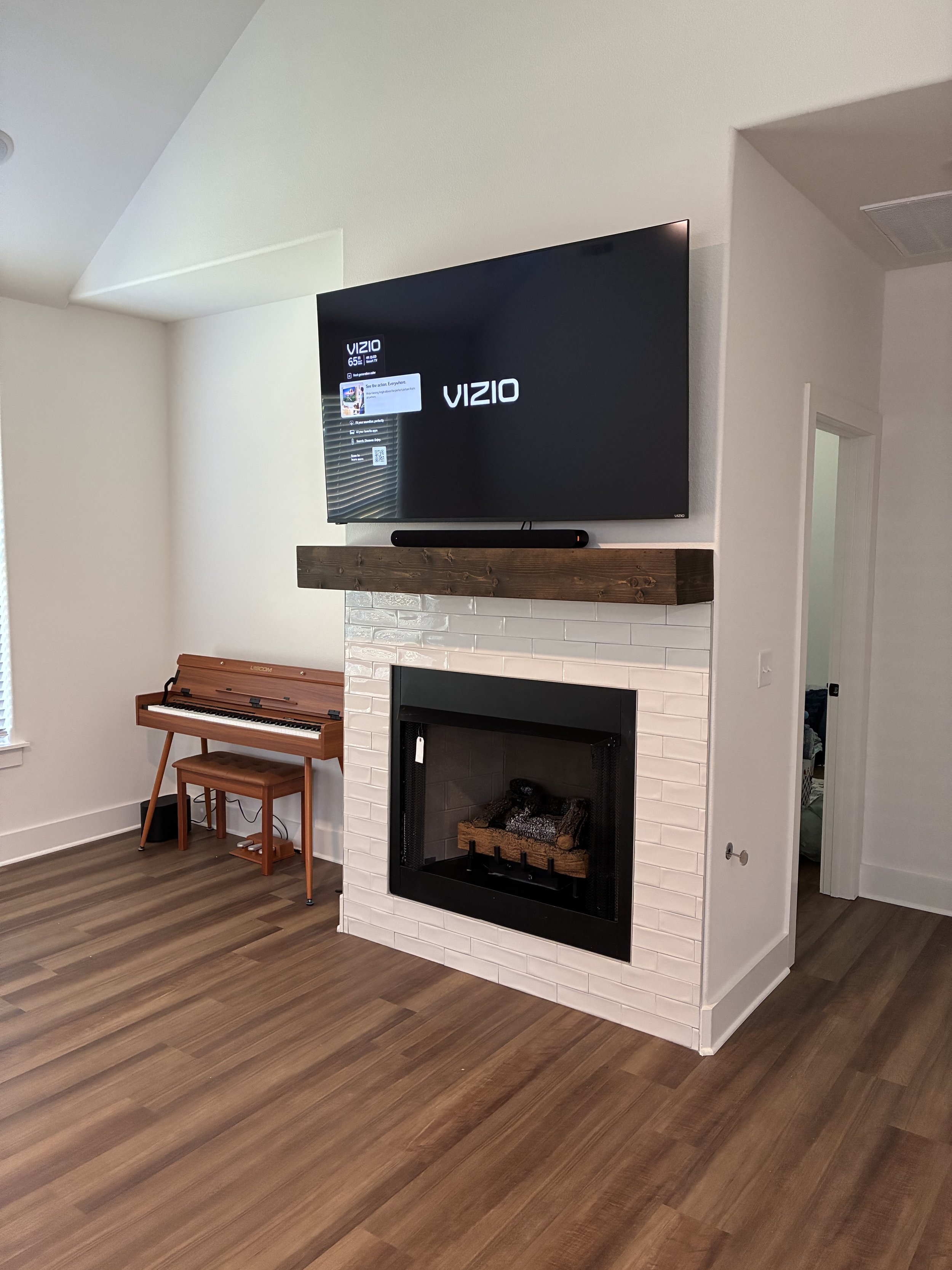 Living room with a mounted flat-screen TV above a white brick fireplace, a wooden mantel, and a small brown piano against the wall. The room has hardwood floors and a partially visible door leading to another room.