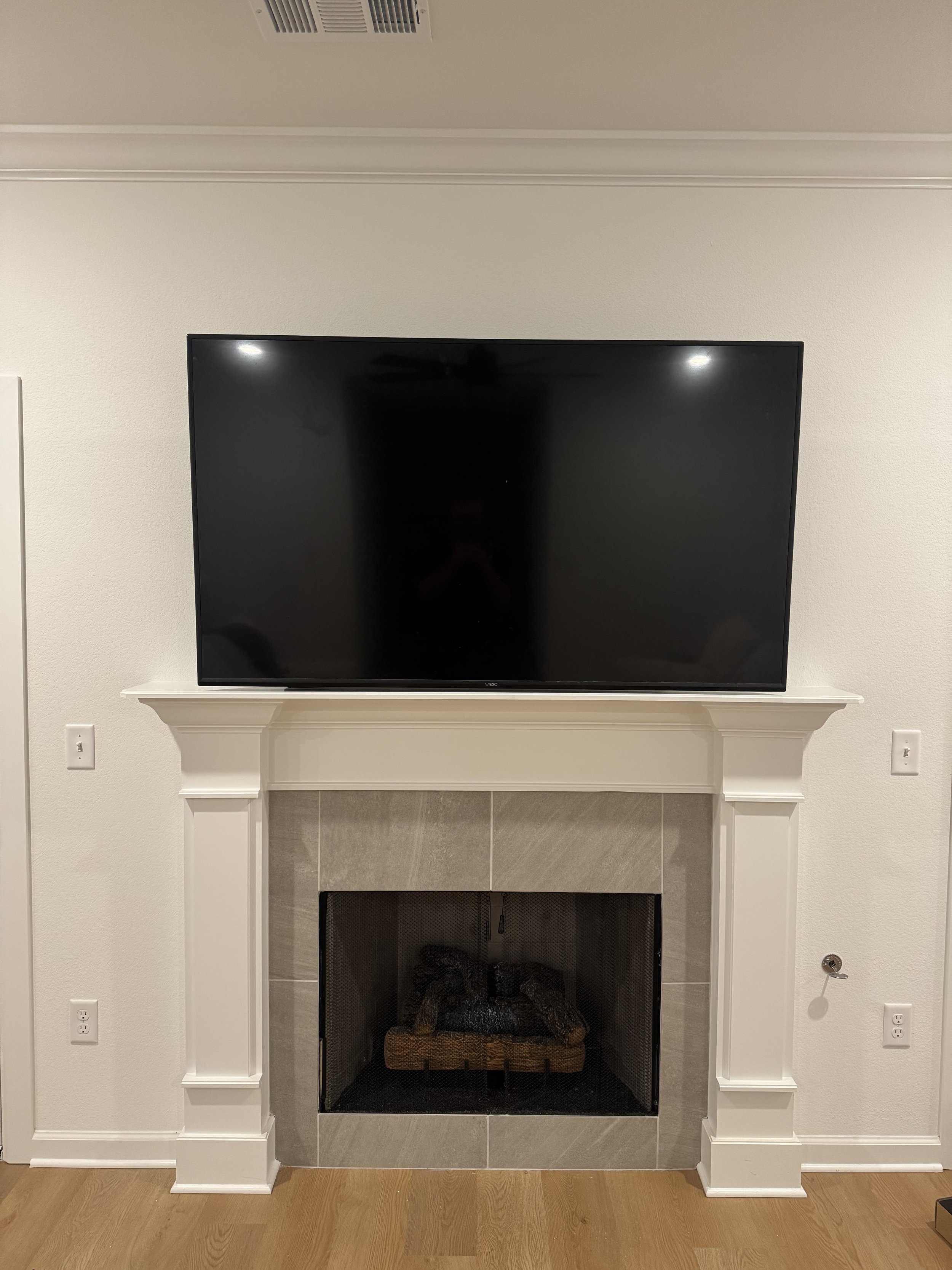 A living room fireplace with a flat-screen TV mounted on the wall above it. The fireplace has a white mantel and a surround of gray tiles, with a decorative log holder inside. The wall is painted white and there are electrical outlets on each side of