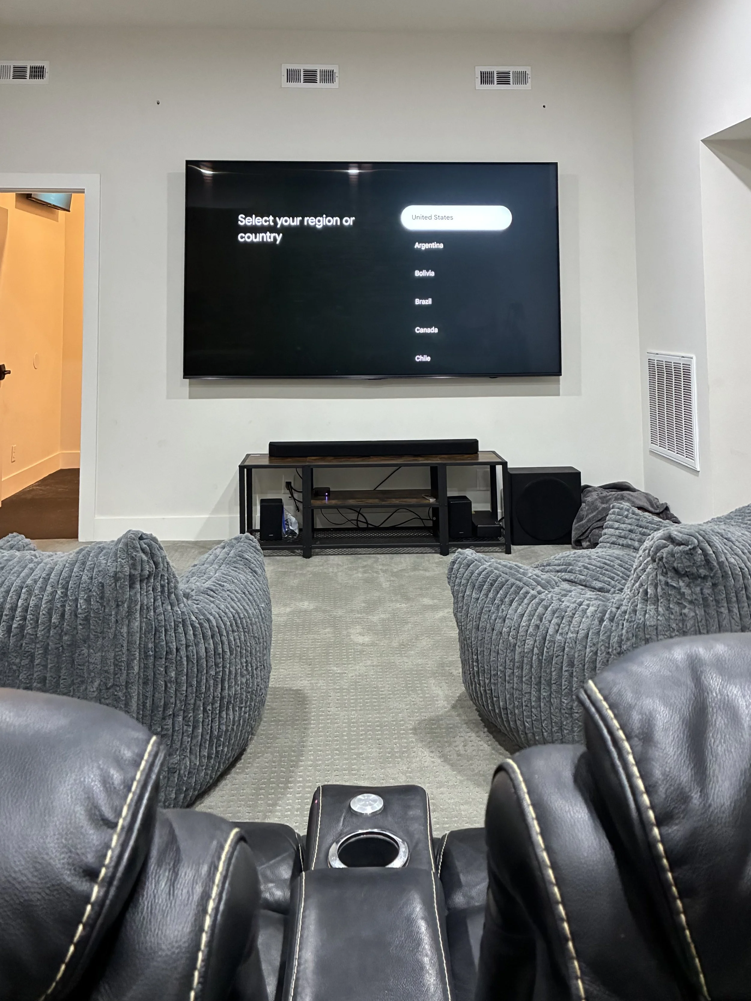 View of a living room with a large flat-screen TV displaying a country selection menu, two gray armchairs, a black leather recliner in the foreground, and a black TV stand with electronic equipment.