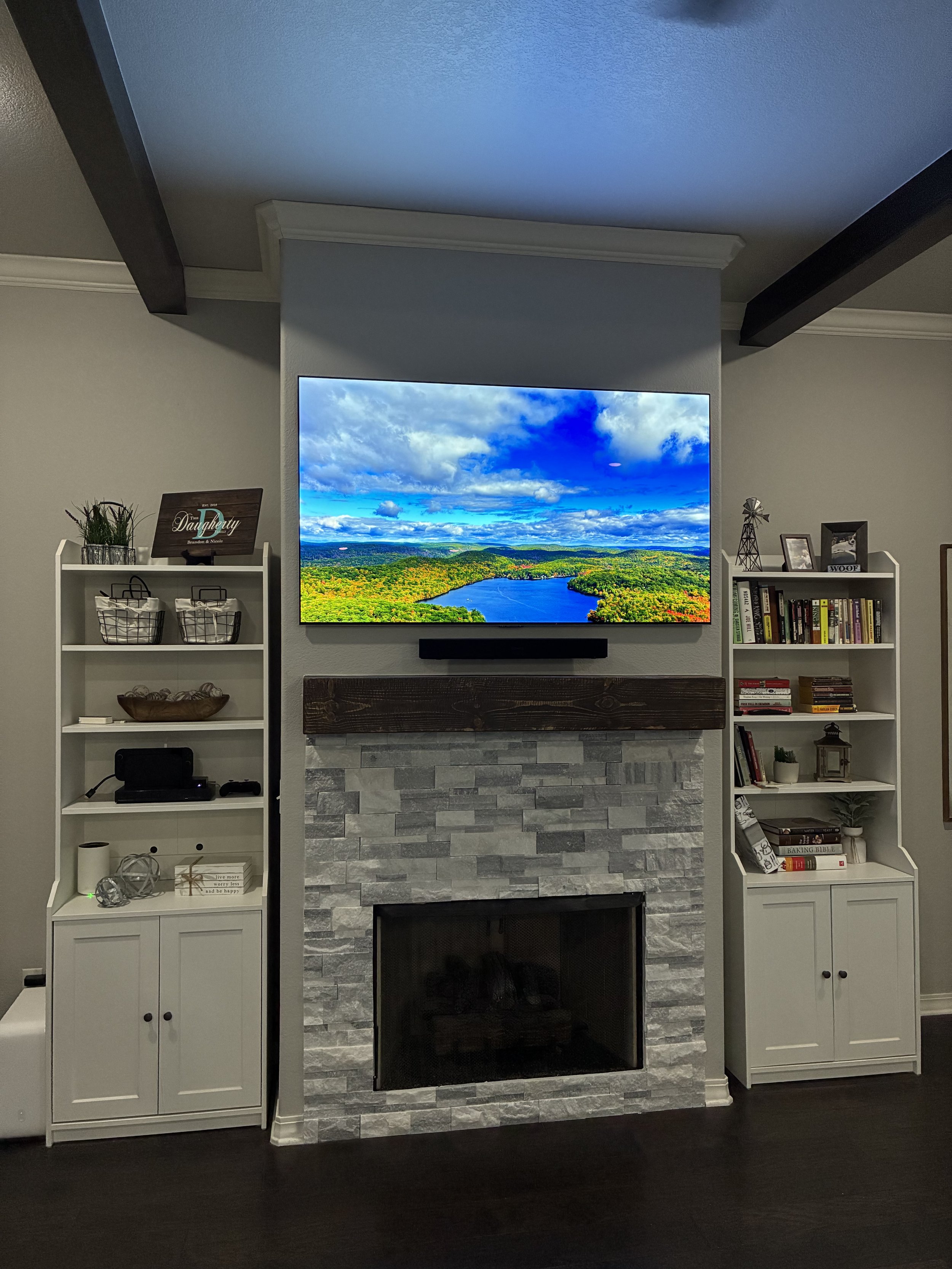 Living room fireplace with mounted flat screen TV displaying a landscape scene, white bookshelves with decorative items on either side of the fireplace, and dark hardwood floors.
