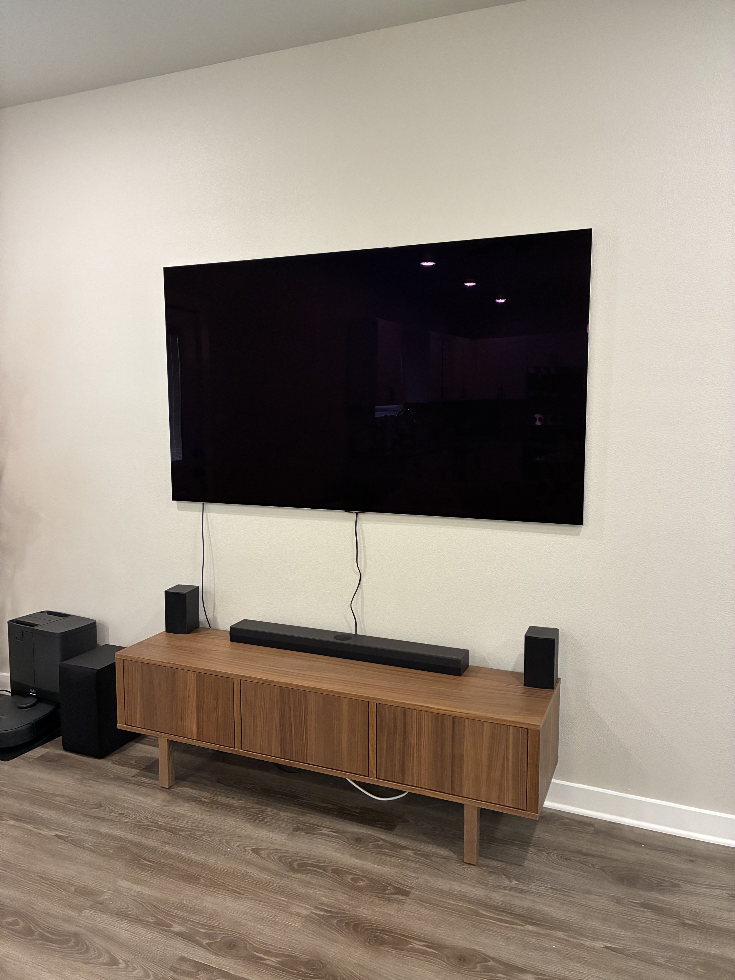 A modern living room with a large flat-screen TV mounted on a white wall, a wooden TV console below, and a soundbar on top of the console. Two small black speakers are positioned on either side of the soundbar. The floor has a wood finish.