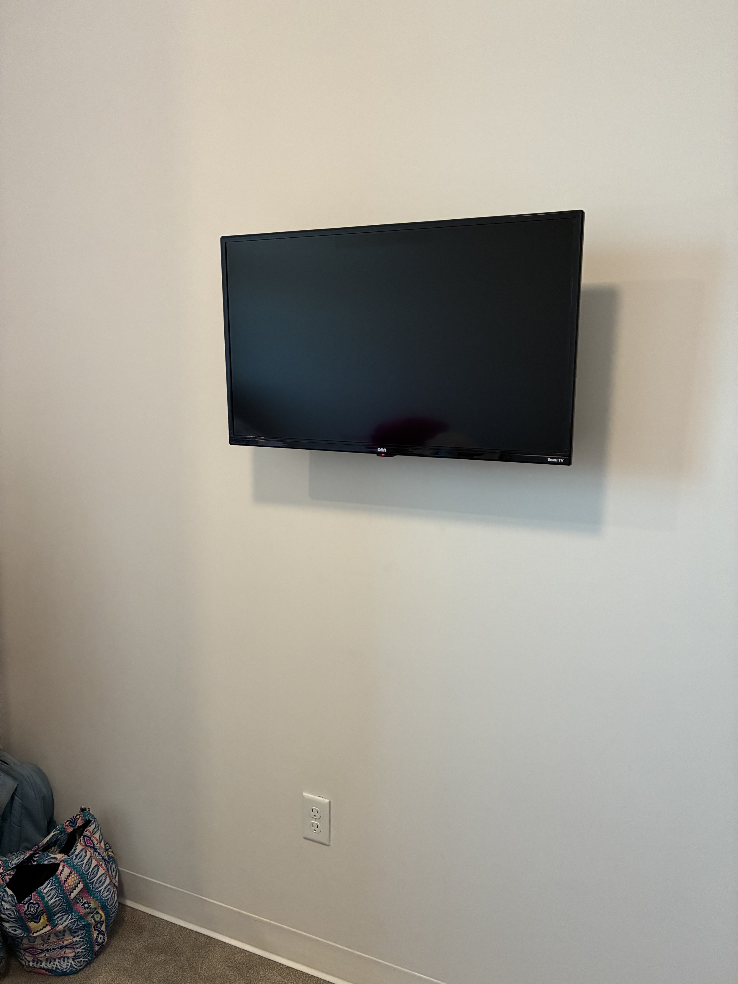 Wall-mounted flat-screen television with a black screen, located in a room with cream-colored walls and carpeted floor. An electrical outlet is visible below the TV, and a bag is on the floor to the left.