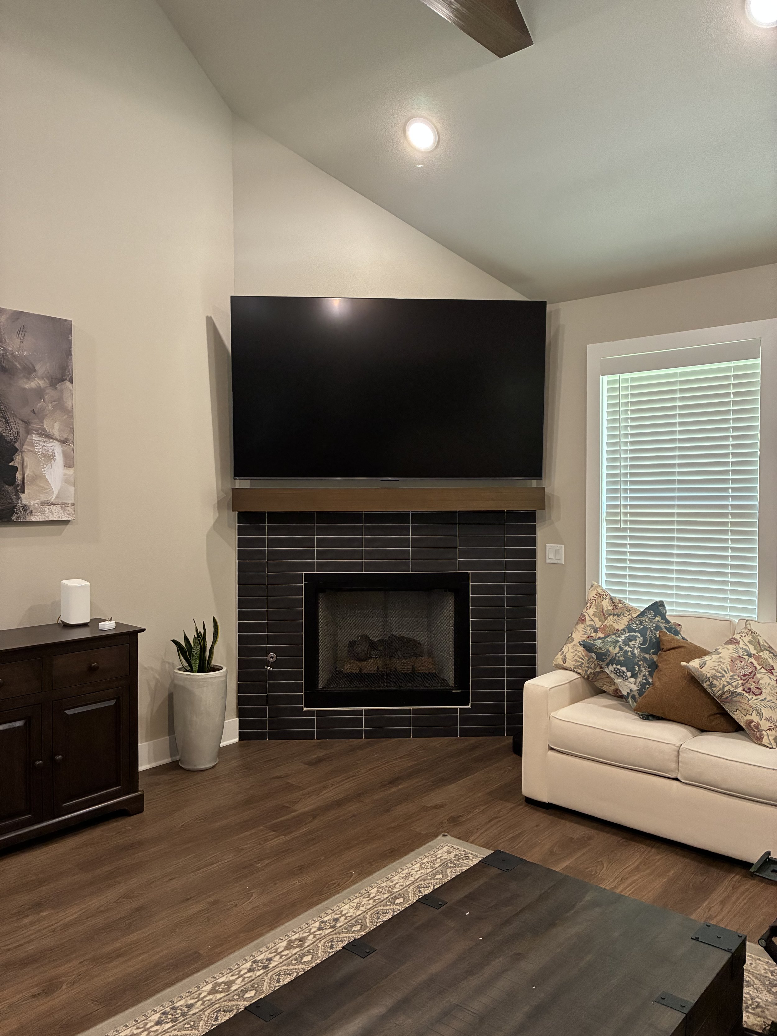 Living room with a black tiled fireplace and a large flat-screen TV mounted above it, a white sofa with decorative pillows, a dark wooden cabinet, a potted plant, and a window with blinds.