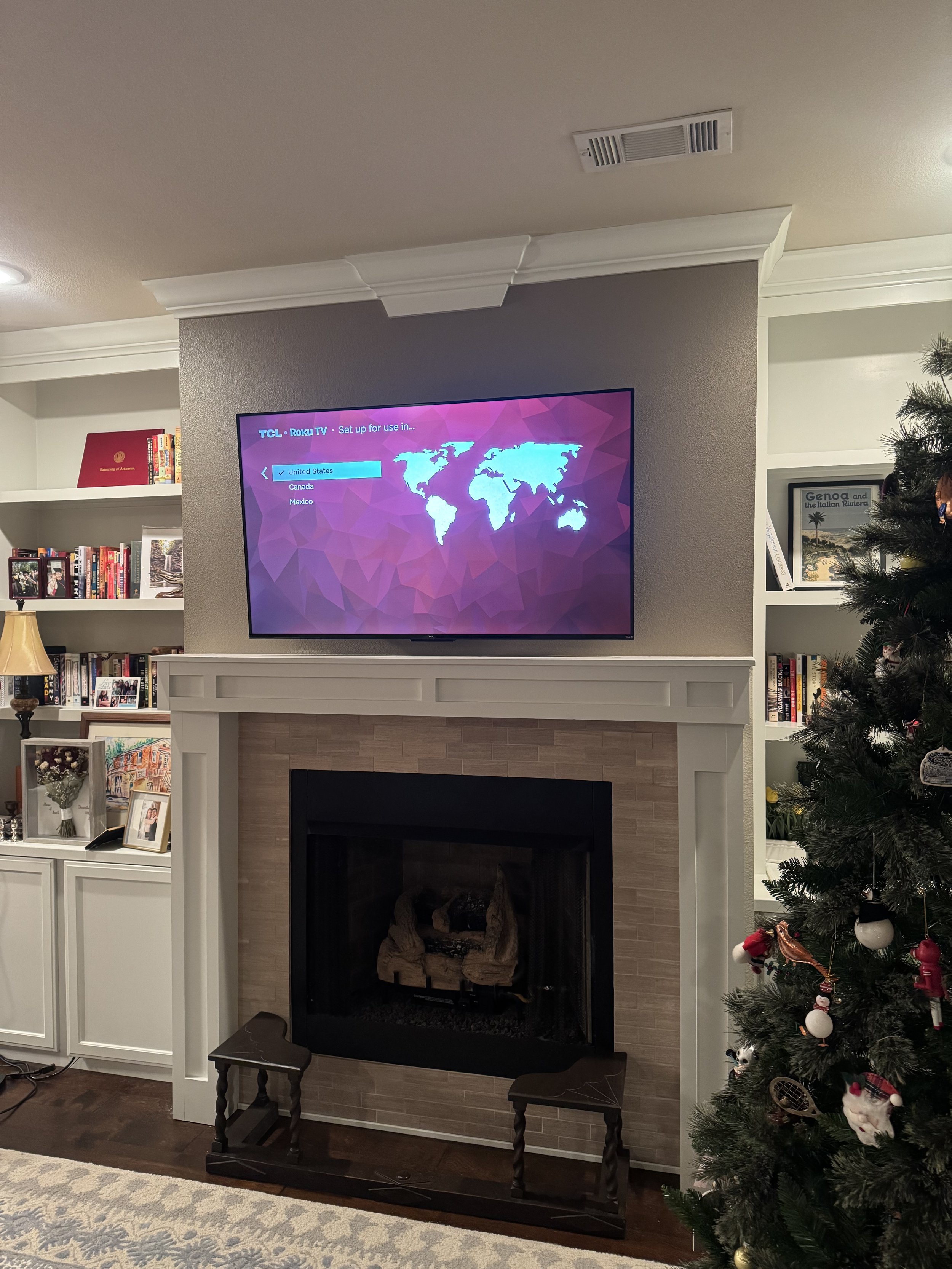 Living room with a fireplace, mounted TV showing a setup screen for the US, Canada, and Mexico, Christmas tree with ornaments on the right, and bookshelves with framed photos and books on the left.