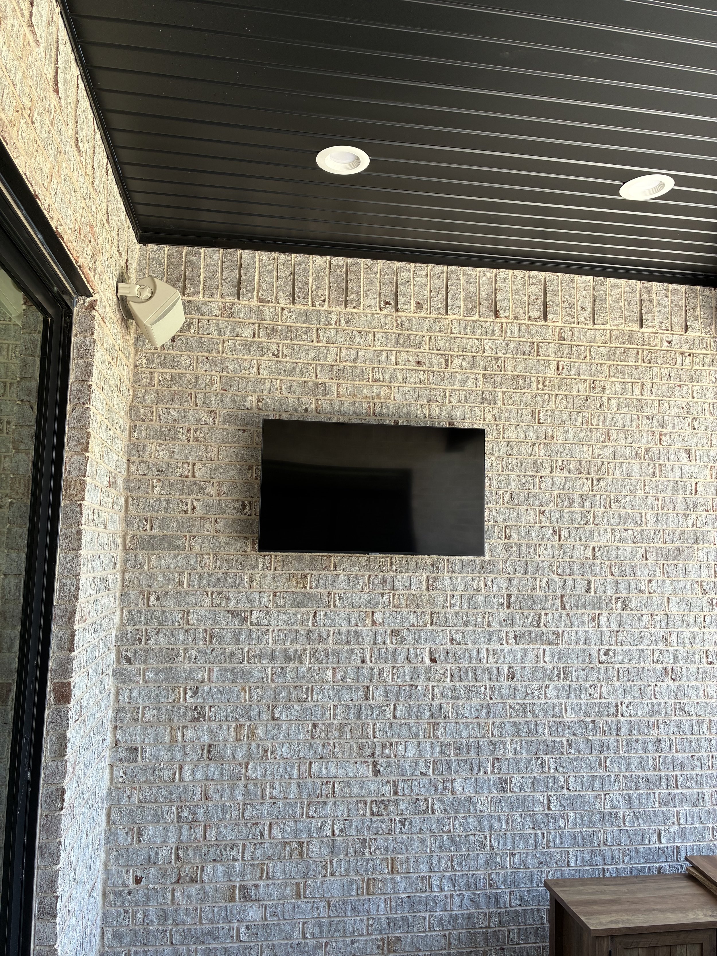 Outdoor patio with brick wall, mounted flat-screen TV, ceiling lights, and a sliding glass door.