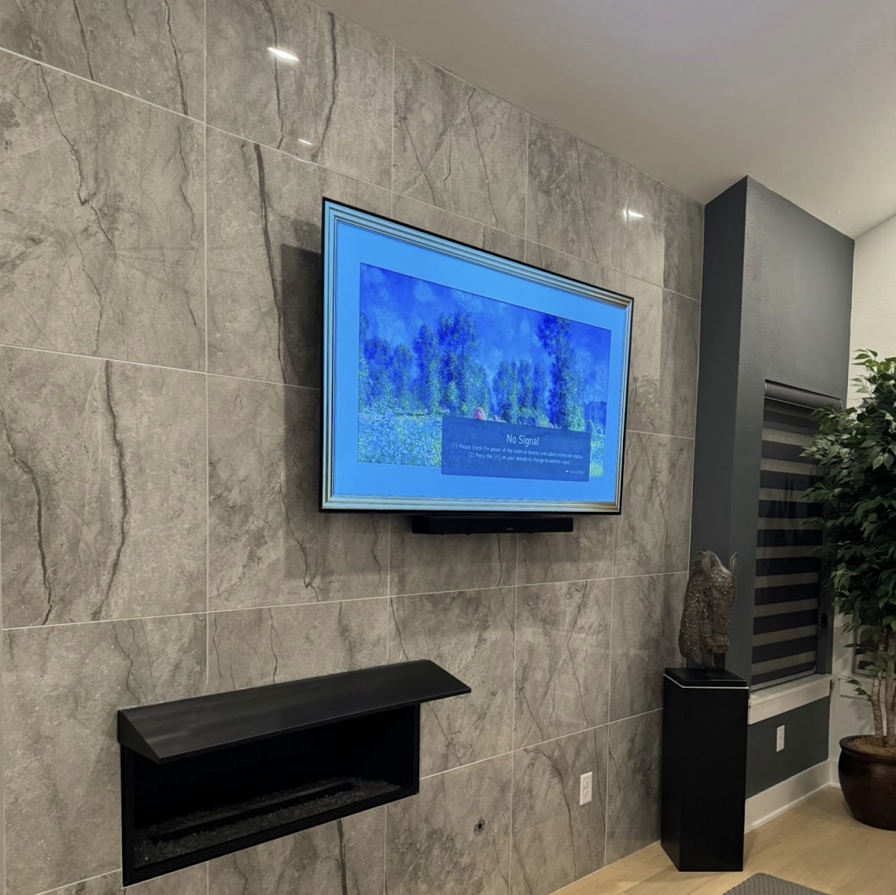 Living room wall with a mounted flat-screen TV displaying a blue screen with a message that says "No Signal". There is a black shelf below the TV, a gray vertical panel with a sculpture on a stand to the right, and a potted plant in the corner.