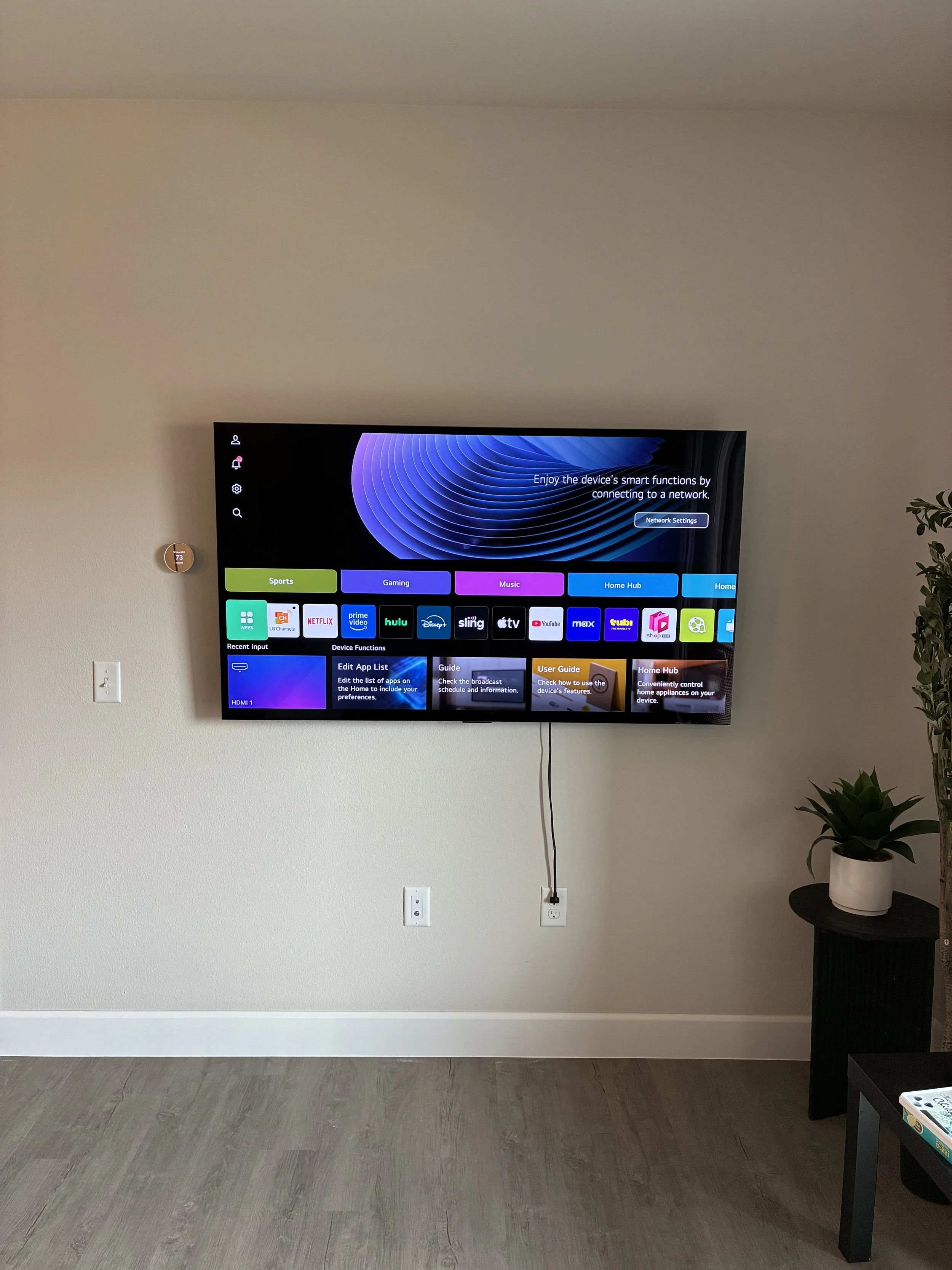 Wall-mounted flat-screen TV displaying smart TV interface with app icons, connected to power outlet via visible cord, next to a small black side table with a potted plant, on a beige wall with a white electrical outlet and a thermostat.